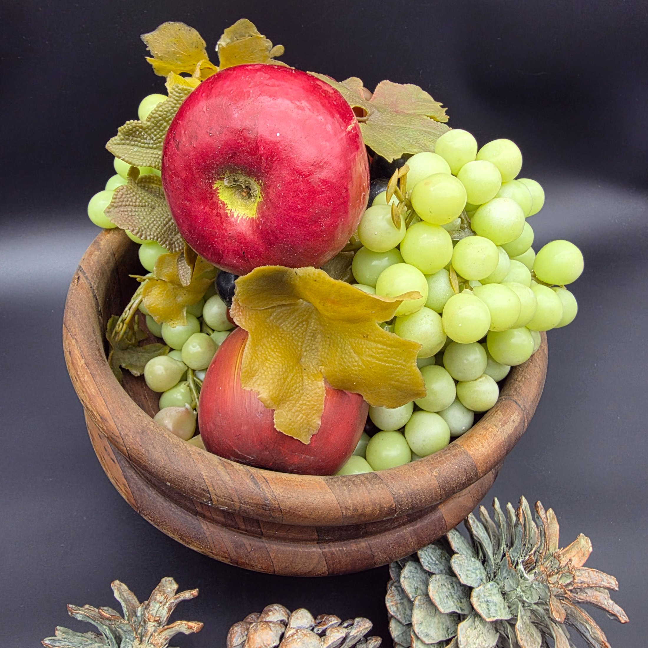 A decorative ensemble featuring a turned wooden bowl filled with artificial fruit (apples and grapes) and faux autumn leaves, accompanied by several large natural pinecones. - View 3