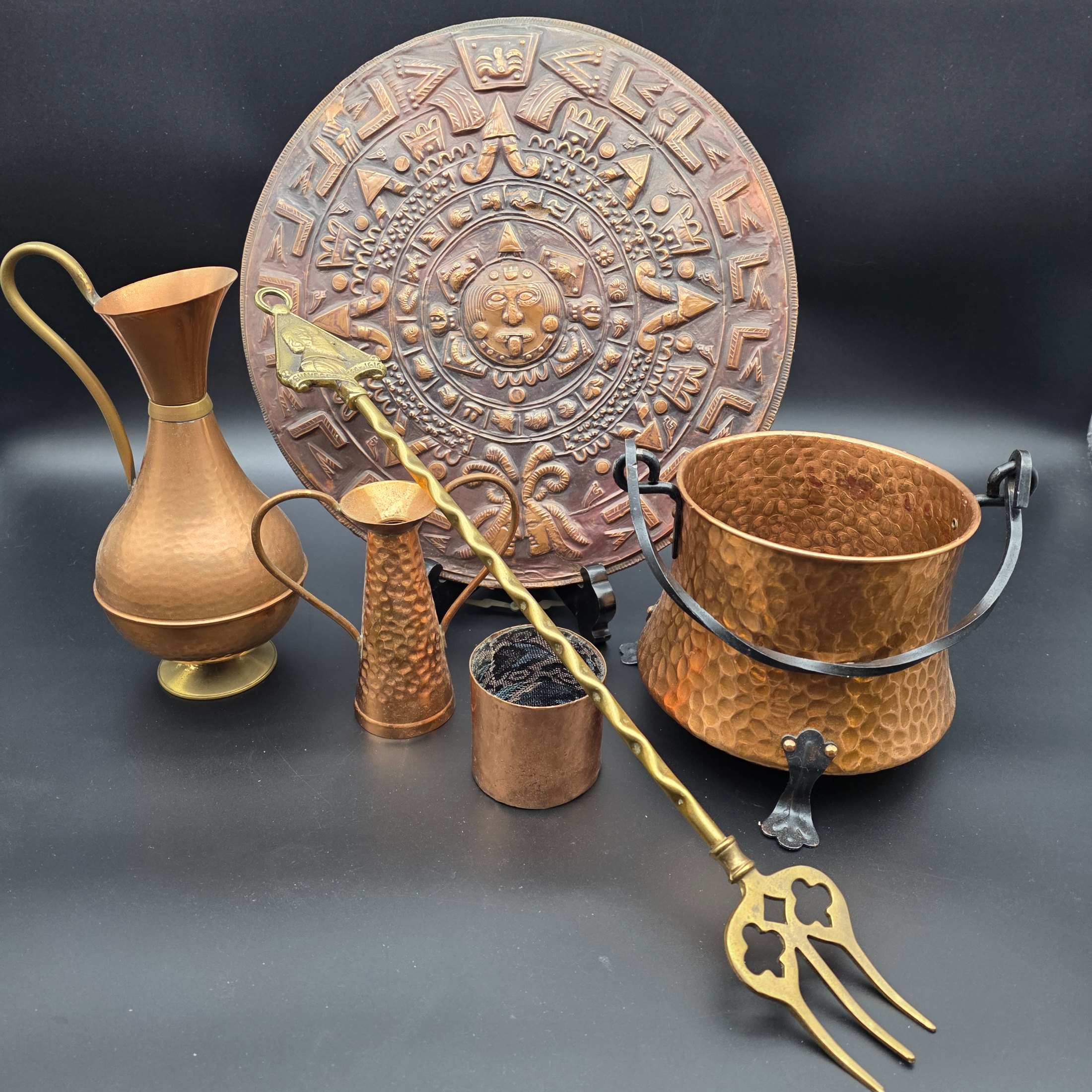 A collection of vintage copper and brass homewares, including an Aztec calendar wall hanging, pitchers, pots, and a Shakespeare-themed toasting fork.