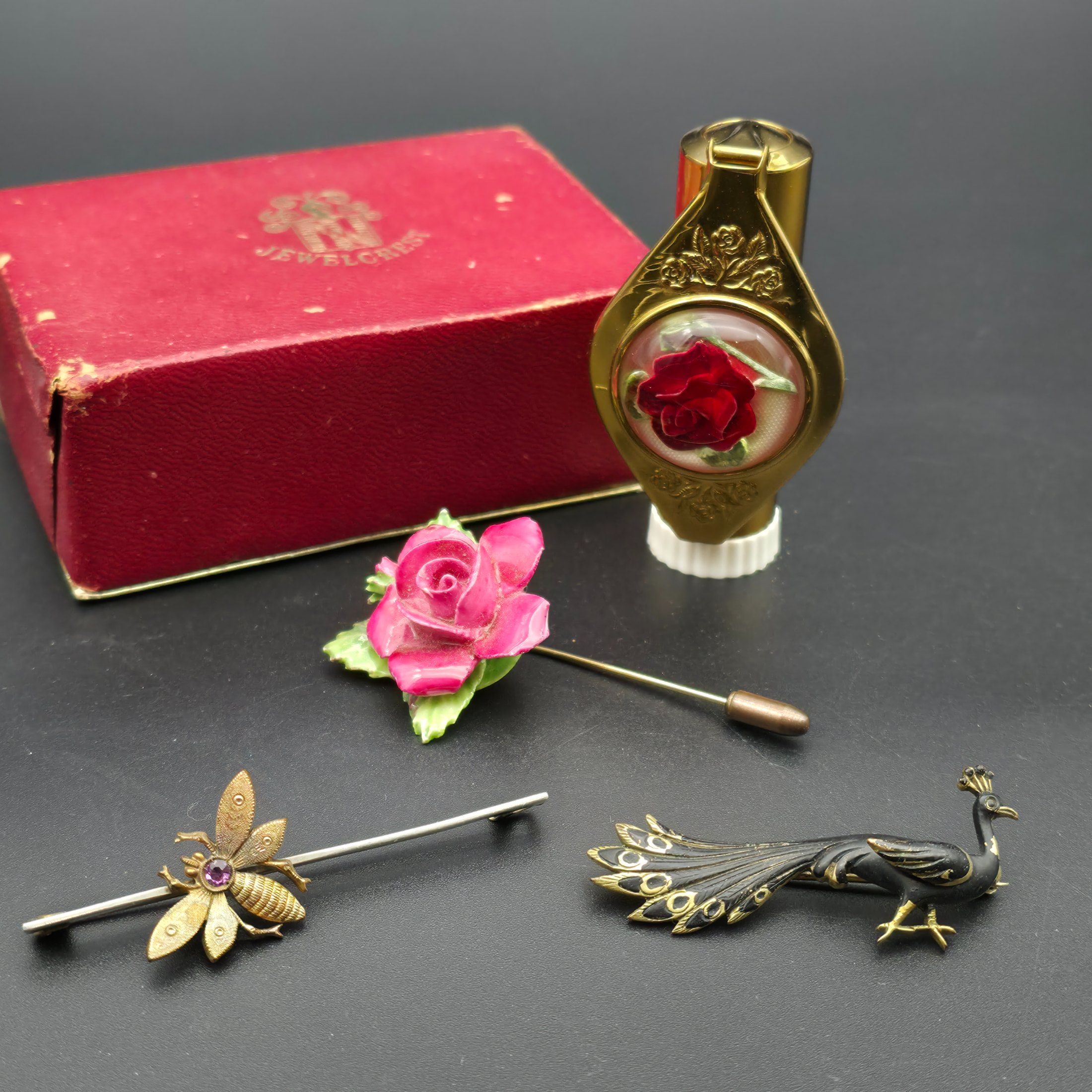 A collection of vintage personal accessories including a bee stick pin, a peacock brooch, a ceramic rose hat pin, and a cased magnifying glass in a red box.