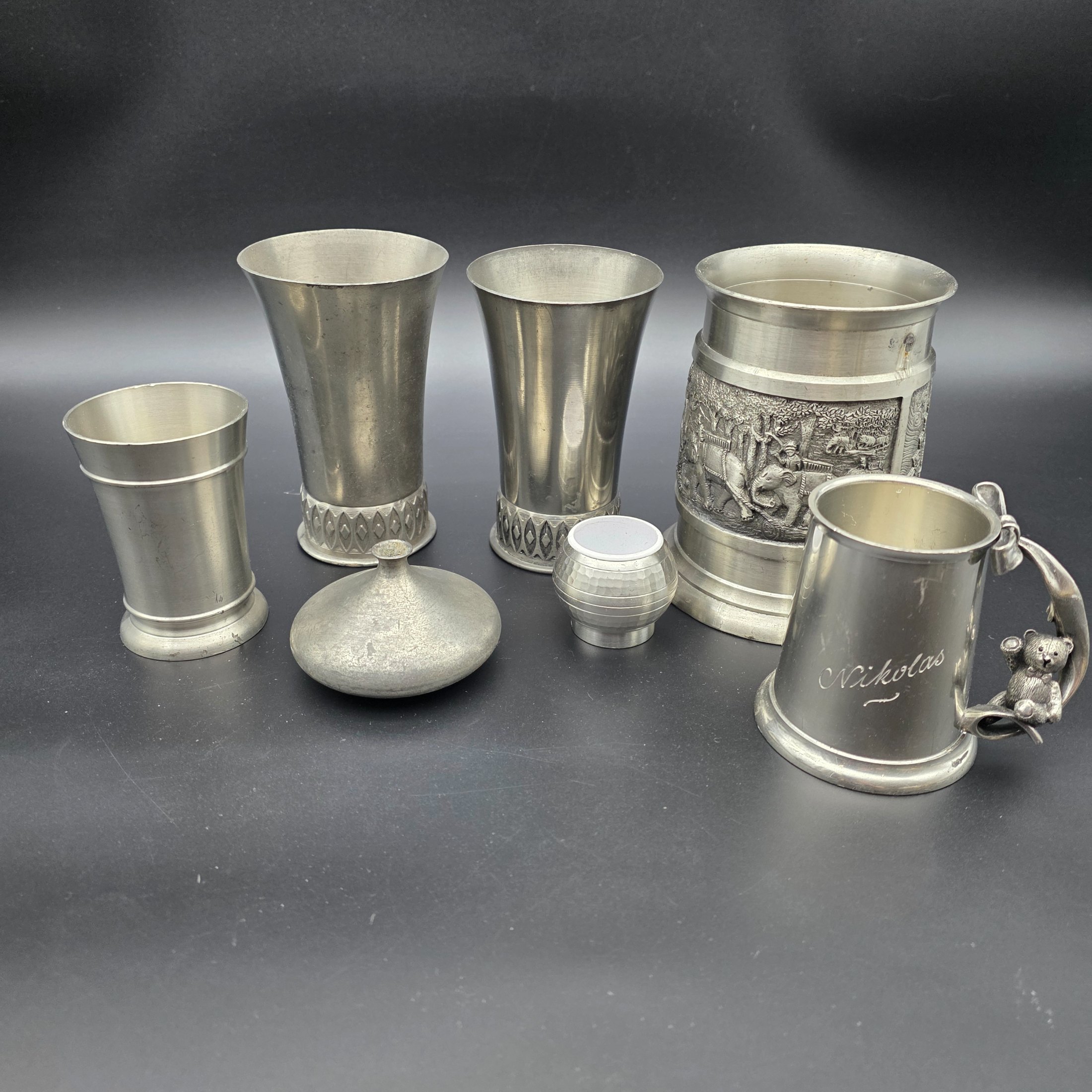 A collection of vintage pewter items including decorative mugs, cups, and small vases, displaying various finishes and makers' marks.