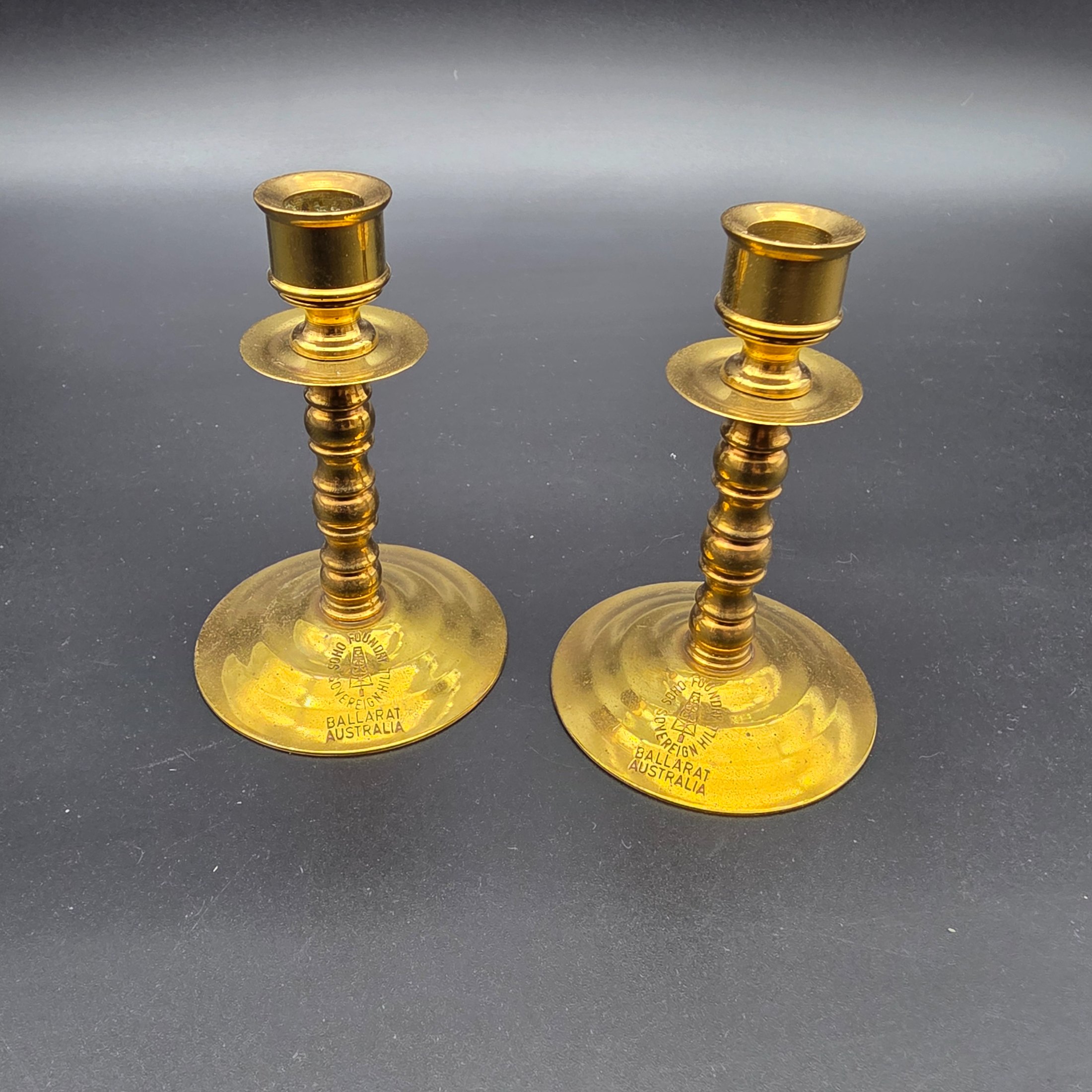 A pair of vintage brass candlesticks, stamped with "Soho Foundry Sovereign Hill Ballarat Australia" and featuring a mining derrick logo, showcasing traditional craftsmanship.