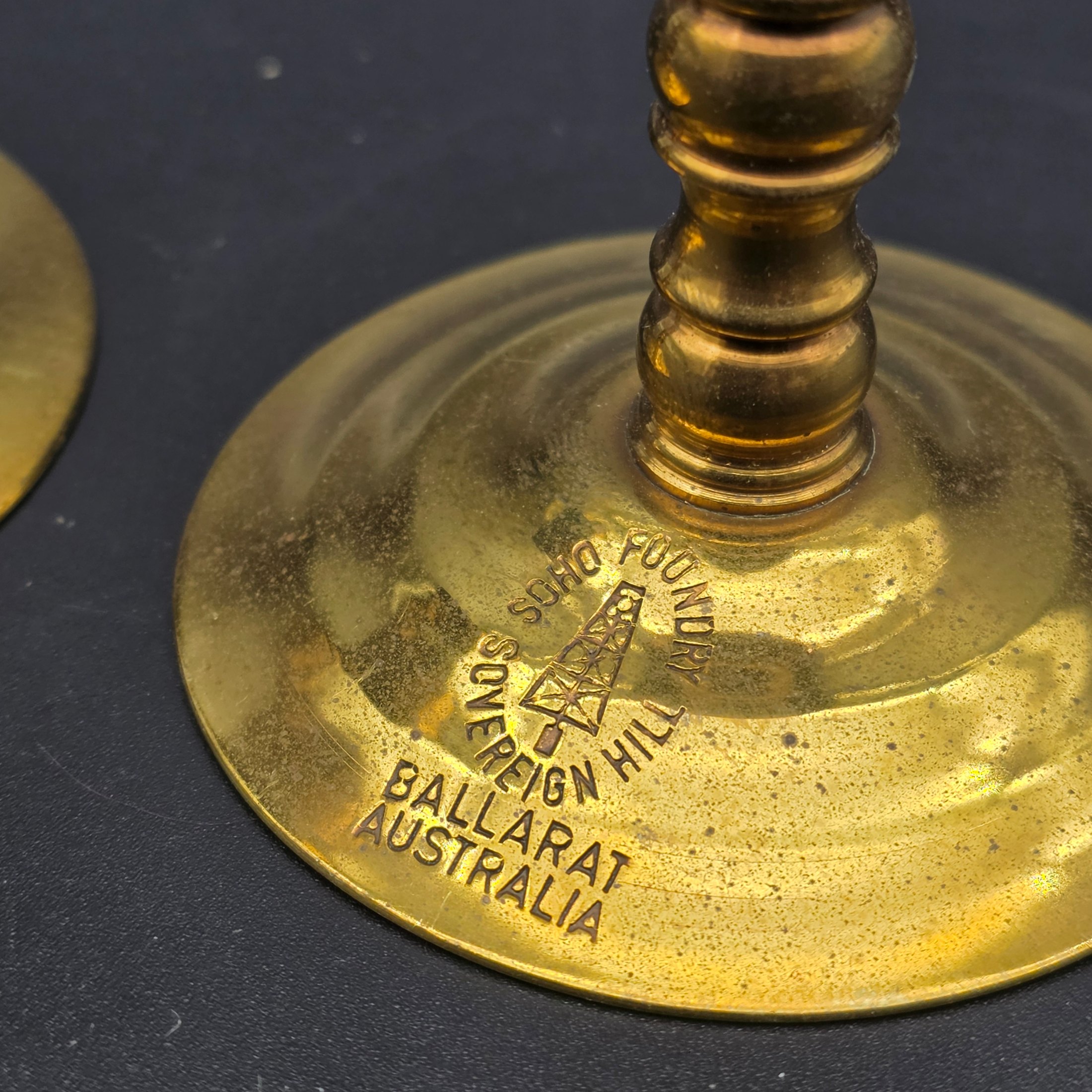 A pair of vintage brass candlesticks, stamped with "Soho Foundry Sovereign Hill Ballarat Australia" and featuring a mining derrick logo, showcasing traditional craftsmanship. - View 2