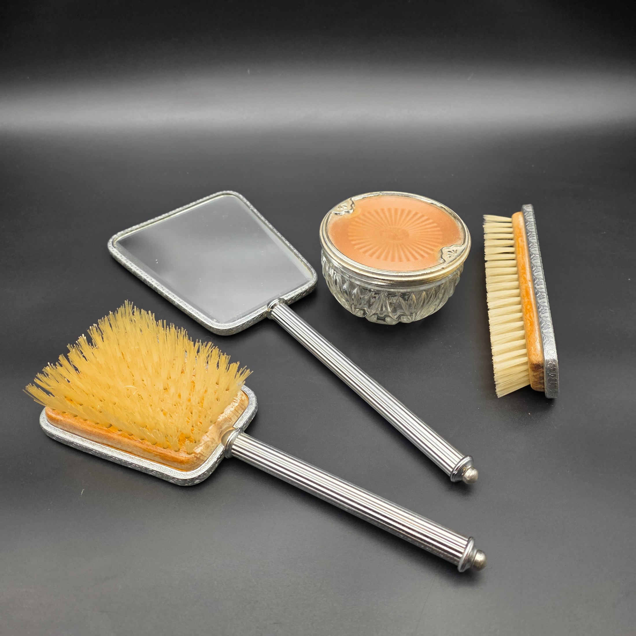 A five-piece vintage Art Deco dressing table vanity set, featuring a hand mirror, two brushes, and a lidded glass powder jar, all with silver-tone metal and rose-gold toned celluloid fan-patterned inserts.