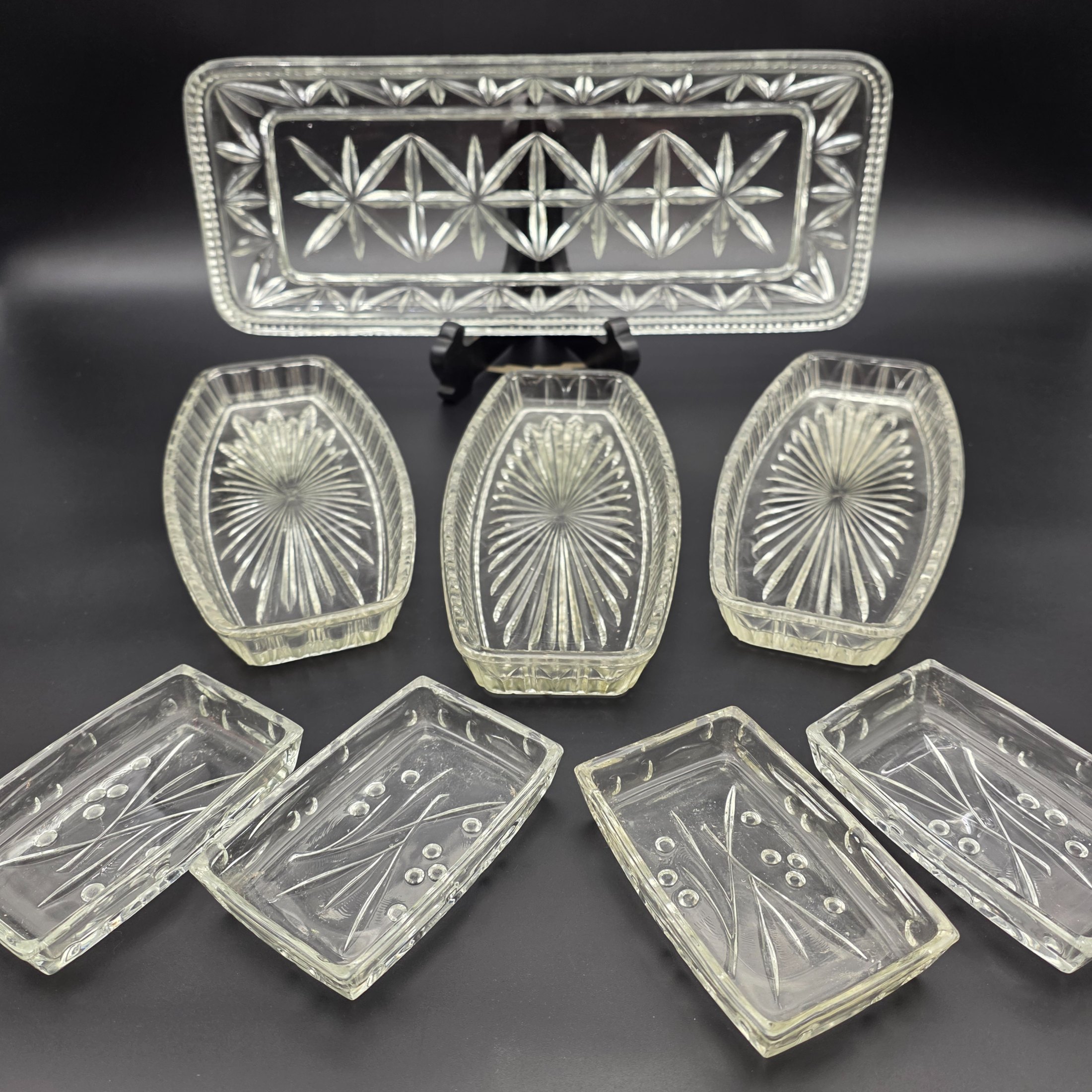 A decorative vintage clear pressed glass serving set, featuring one large rectangular tray, three oval dishes, and four smaller rectangular dishes, showcasing intricate pressed patterns.