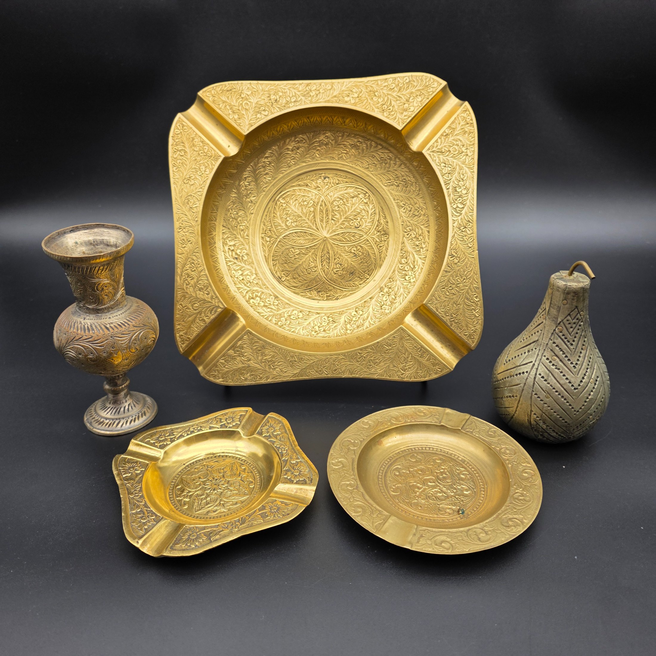 A collection of five vintage engraved brass and bronze decorative items, including three ashtrays, a vase, and a pear-shaped container.