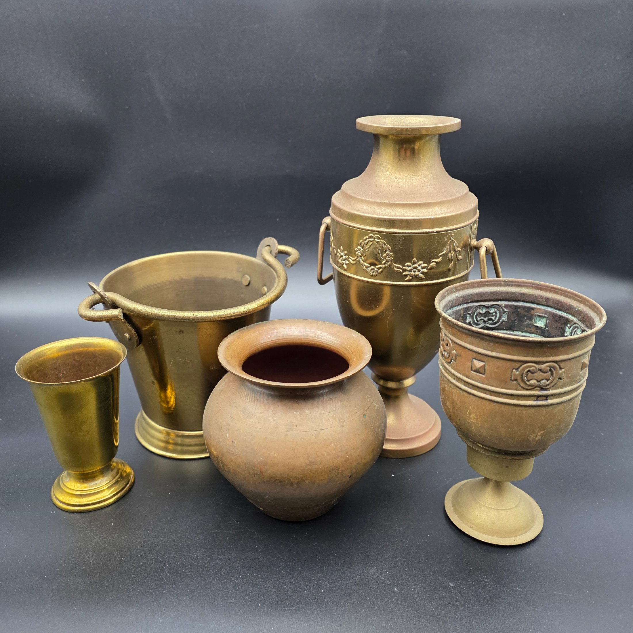 A varied collection of five vintage decorative vessels crafted from brass and copper, including a small footed goblet, a rounded copper pot, a brass bucket with a handle, a tall handled brass urn, and a footed brass goblet with ornate banding. At least one piece bears a 'MADE IN INDIA' label, indicating a probable origin for the entire group.
