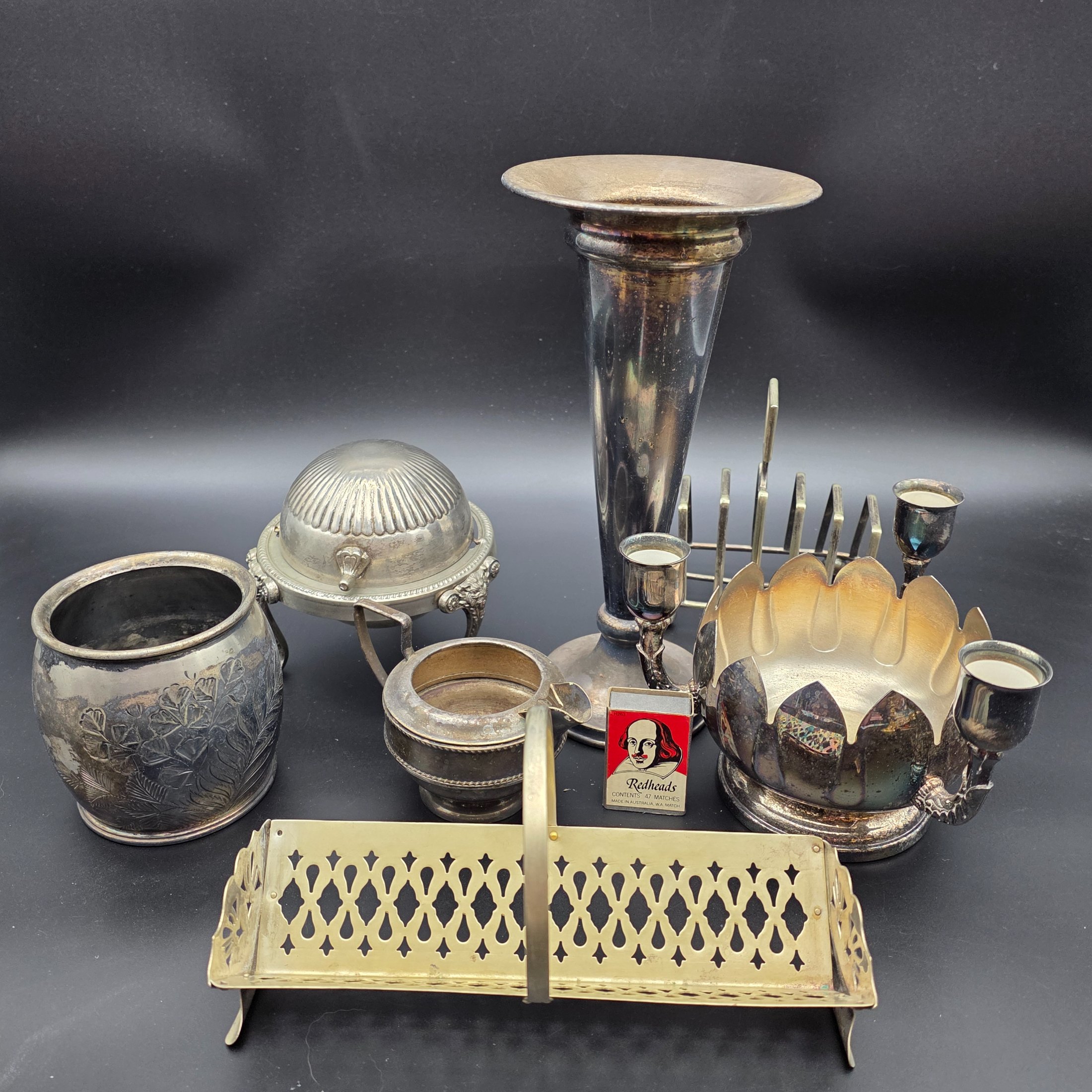 A varied collection of vintage silver-plated items including a butter dish, vase, creamer, bowls, and a toast rack, showing signs of heavy tarnishing and wear.