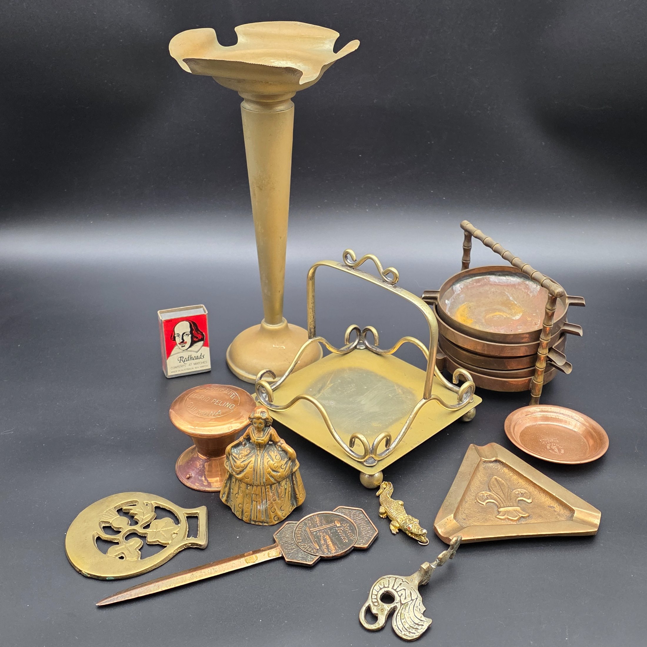 A diverse collection of vintage decorative items crafted from brass and copper, including a tall vase, various ashtrays, a small handled tray, a bell, a letter opener, and other small metallic objects, likely from the mid-20th century.