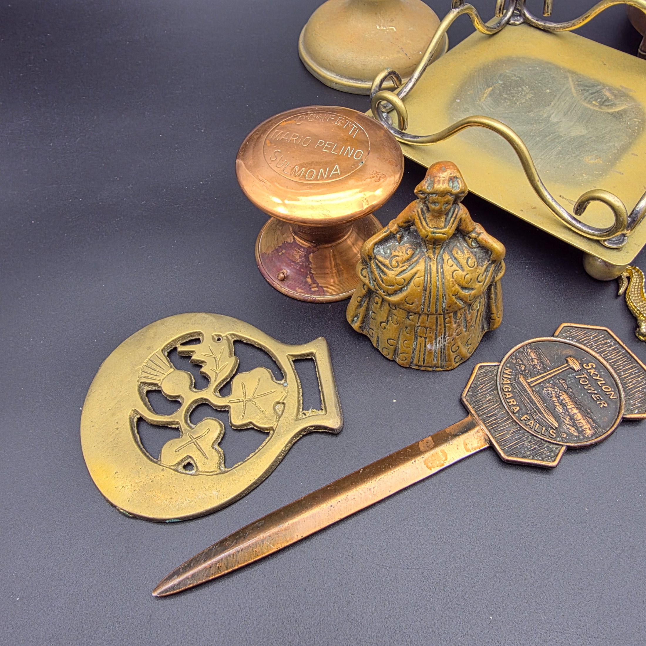 A diverse collection of vintage decorative items crafted from brass and copper, including a tall vase, various ashtrays, a small handled tray, a bell, a letter opener, and other small metallic objects, likely from the mid-20th century. - View 2