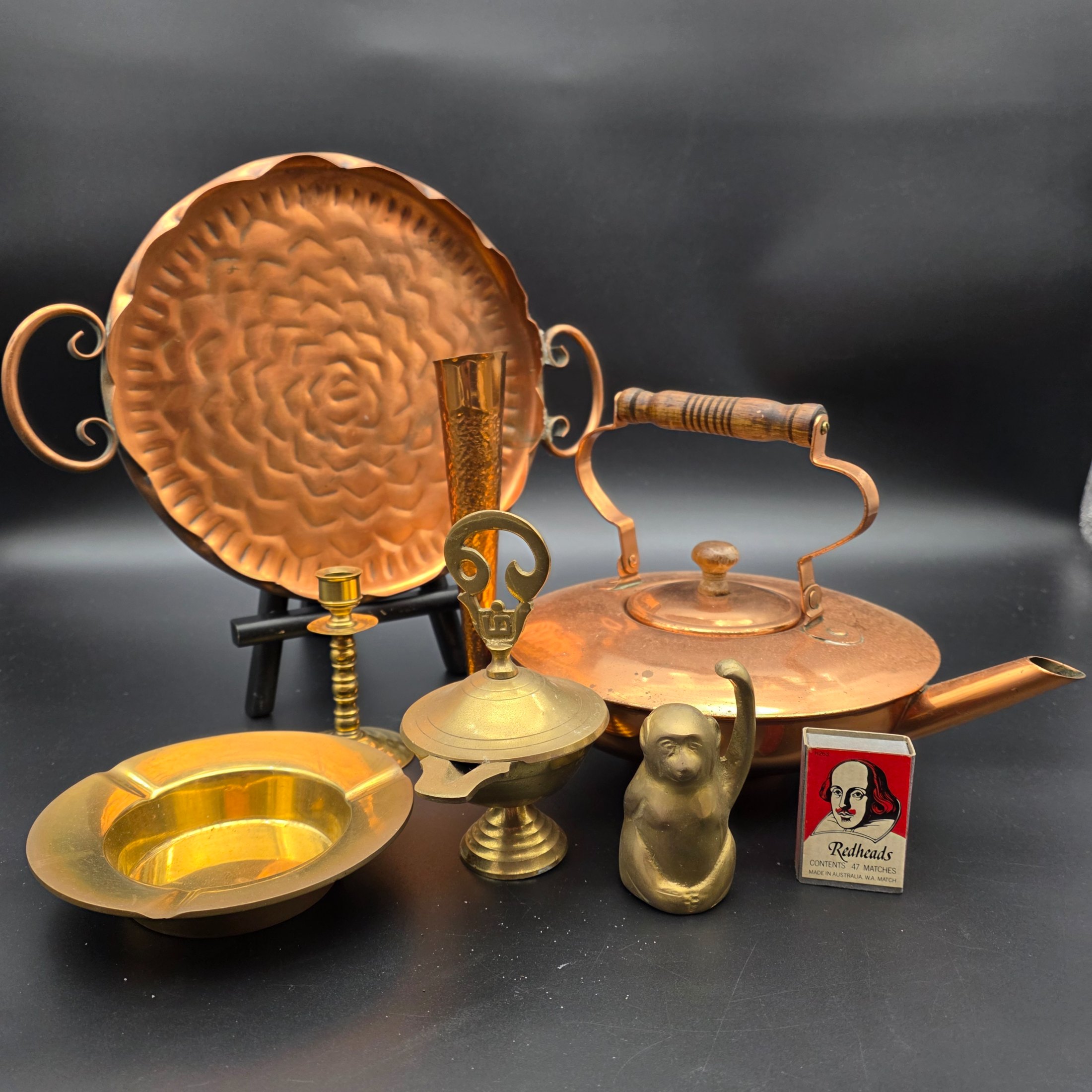 A collection of vintage copper and brass homewares, featuring a large decorative tray, a kettle, a candlestick, an ashtray, a lidded pot, a small vase, and a monkey bell.