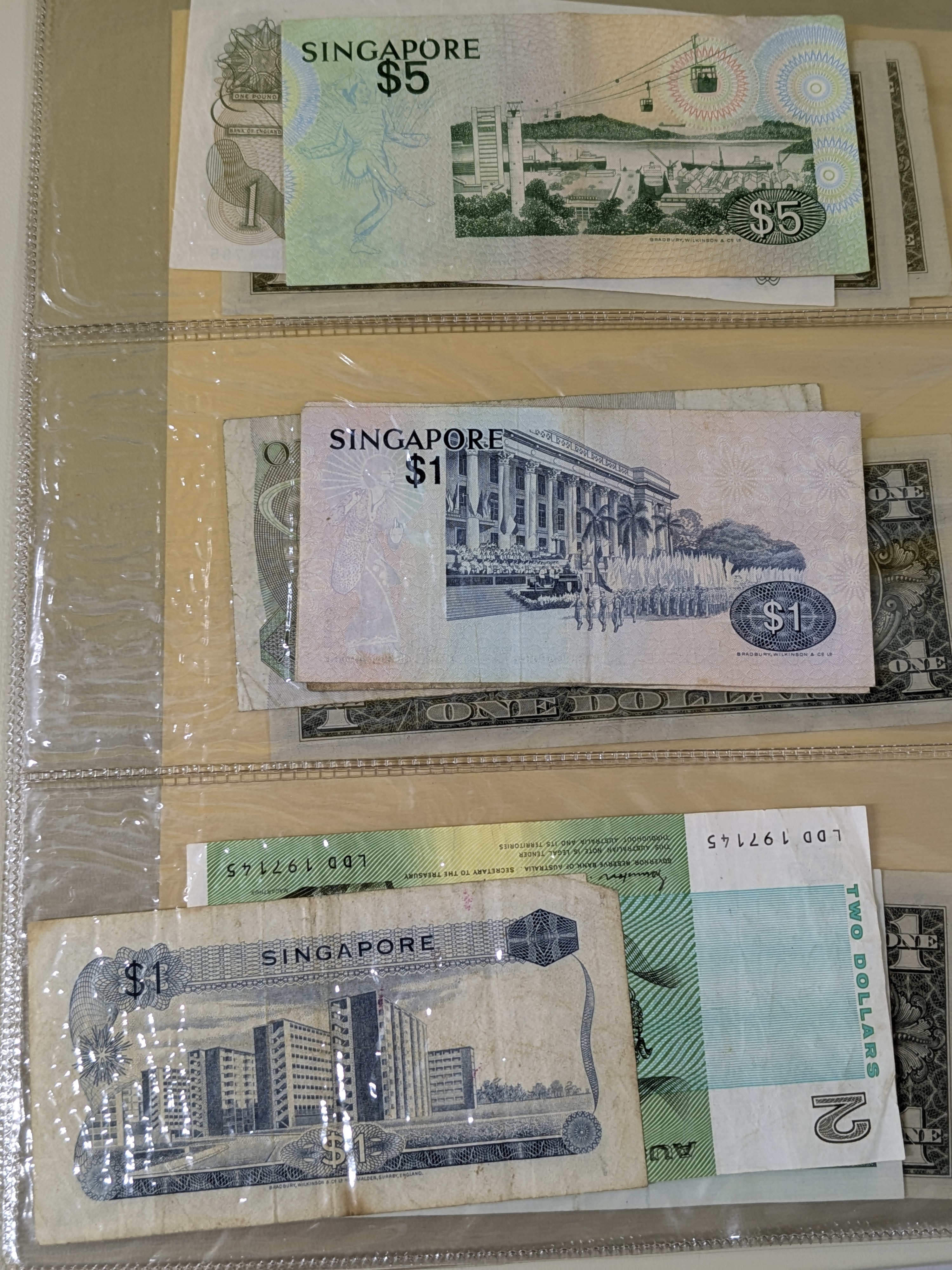 A collection of international banknotes housed within a dark blue 'FIRST DAY COVERS' album, featuring currency from Singapore, Australia, the United Kingdom, and the United States, dating from various periods. - View 2