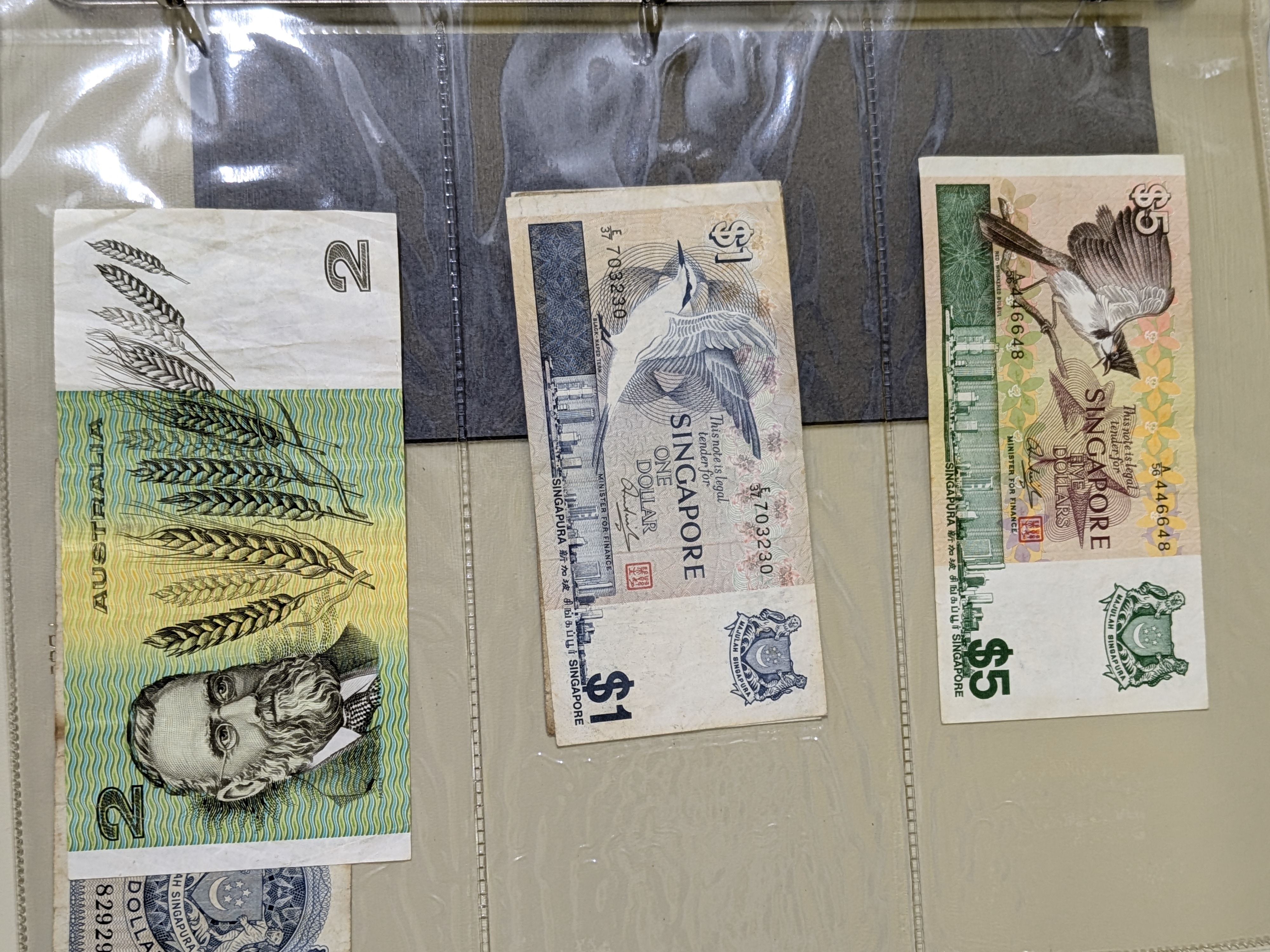 A collection of international banknotes housed within a dark blue 'FIRST DAY COVERS' album, featuring currency from Singapore, Australia, the United Kingdom, and the United States, dating from various periods. - View 6