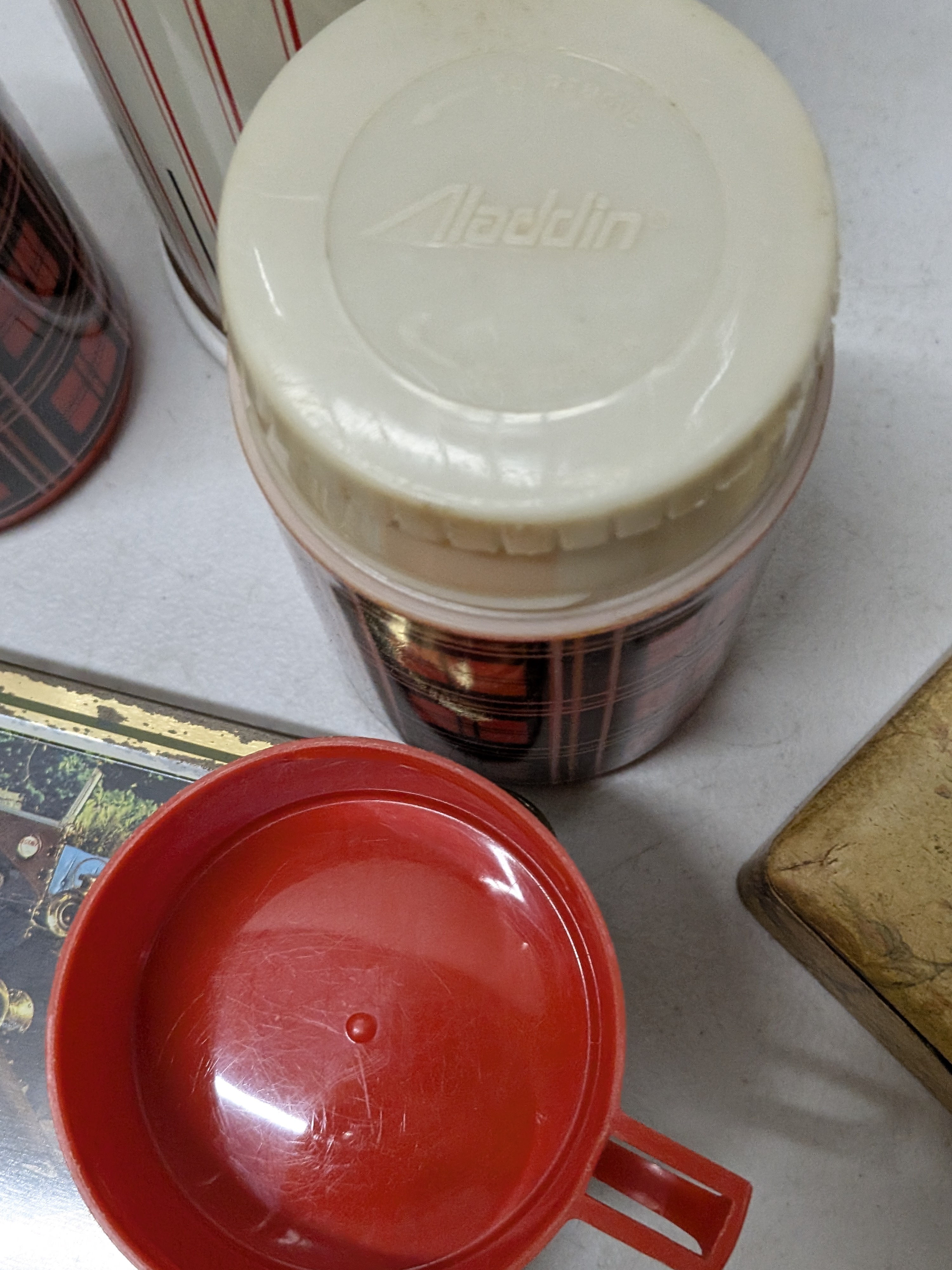 A charming collection of vintage homewares featuring a large white Thermos Midi flask, two smaller red plaid Aladdin thermal bottles, a rectangular metal tin adorned with a classic car image, and a small hinged wooden box with a floral motif. - View 3