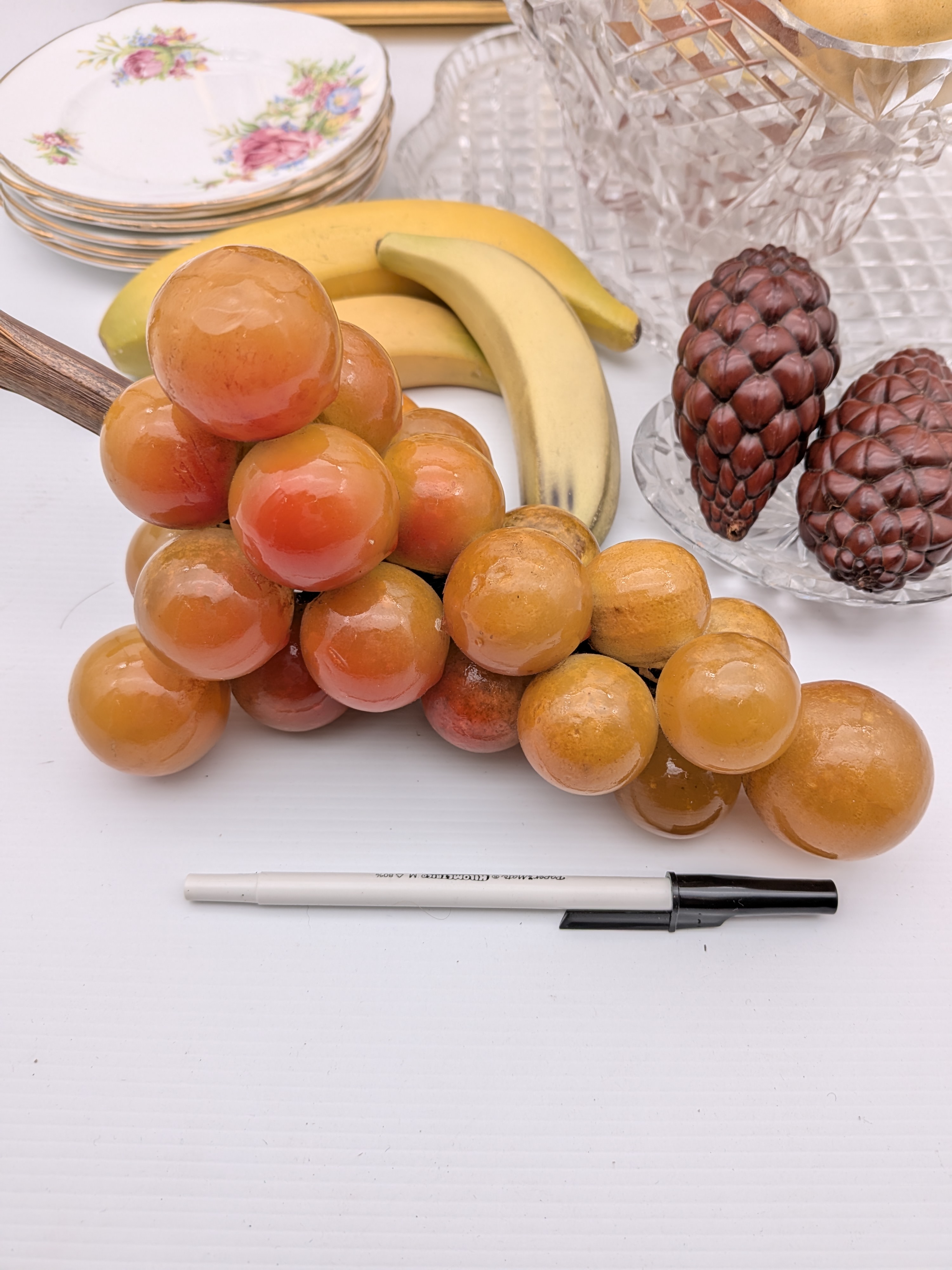 A diverse collection of vintage decorative household sundries, featuring a framed bird relief art piece, a cut-crystal fruit bowl with faux fruit, a stack of ceramic floral side plates, and additional faux fruit. - View 2