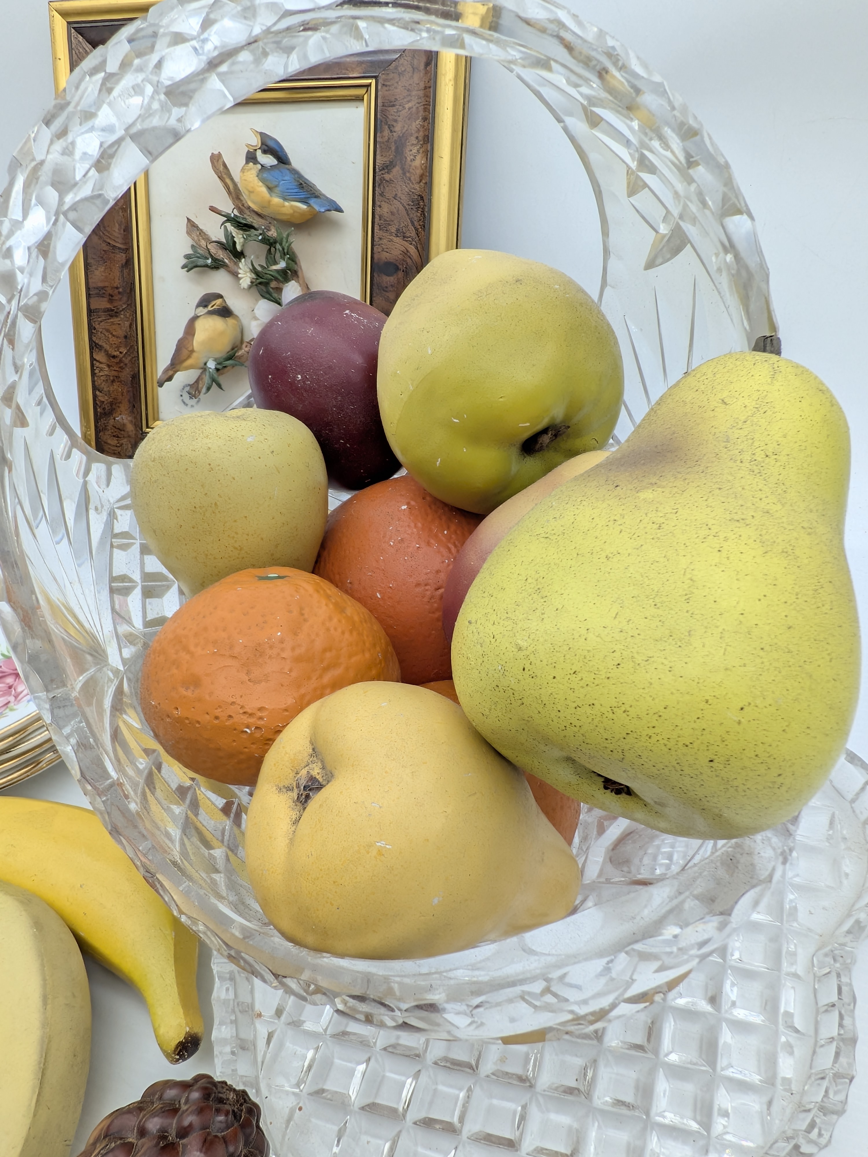 A diverse collection of vintage decorative household sundries, featuring a framed bird relief art piece, a cut-crystal fruit bowl with faux fruit, a stack of ceramic floral side plates, and additional faux fruit. - View 4