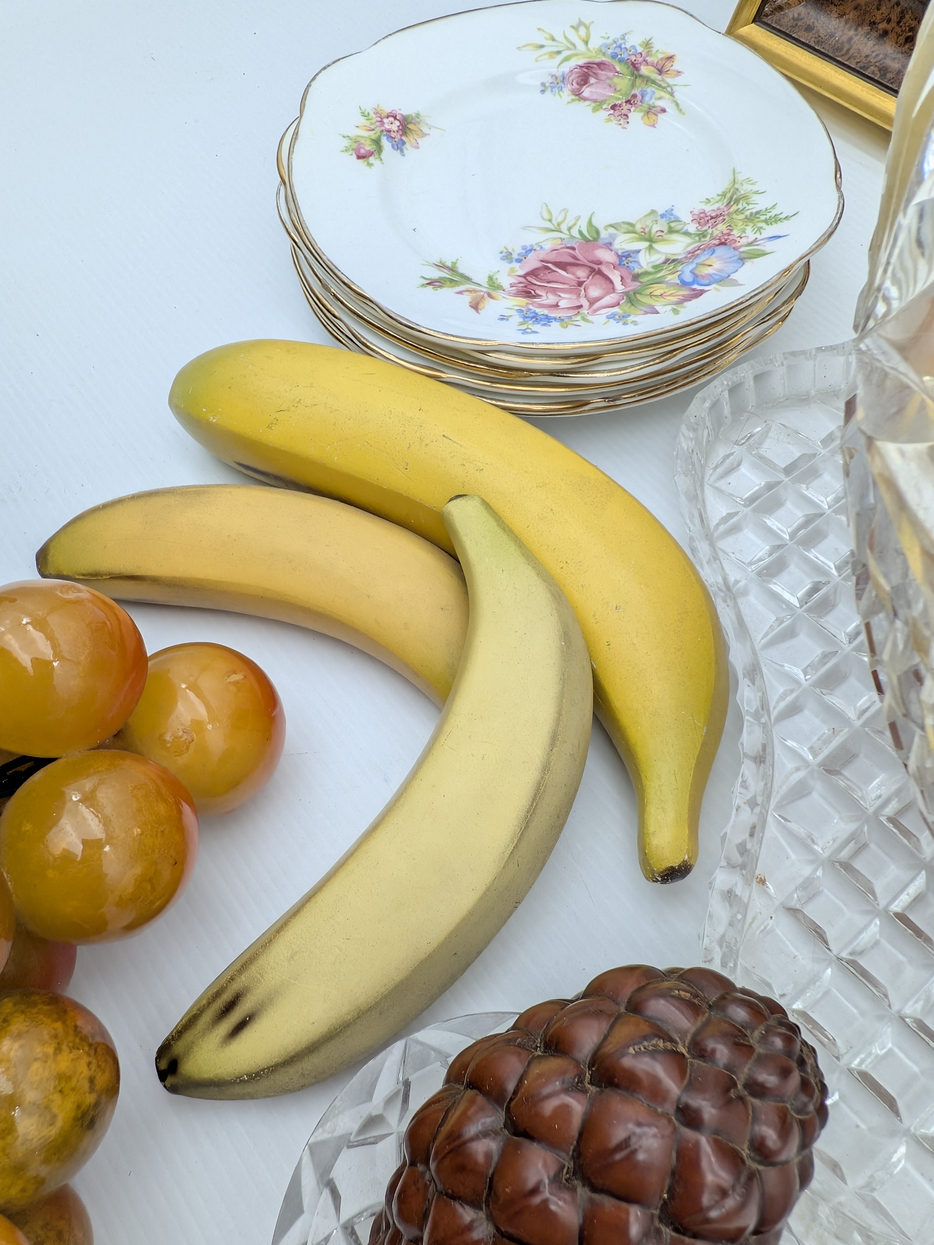 A diverse collection of vintage decorative household sundries, featuring a framed bird relief art piece, a cut-crystal fruit bowl with faux fruit, a stack of ceramic floral side plates, and additional faux fruit. - View 5