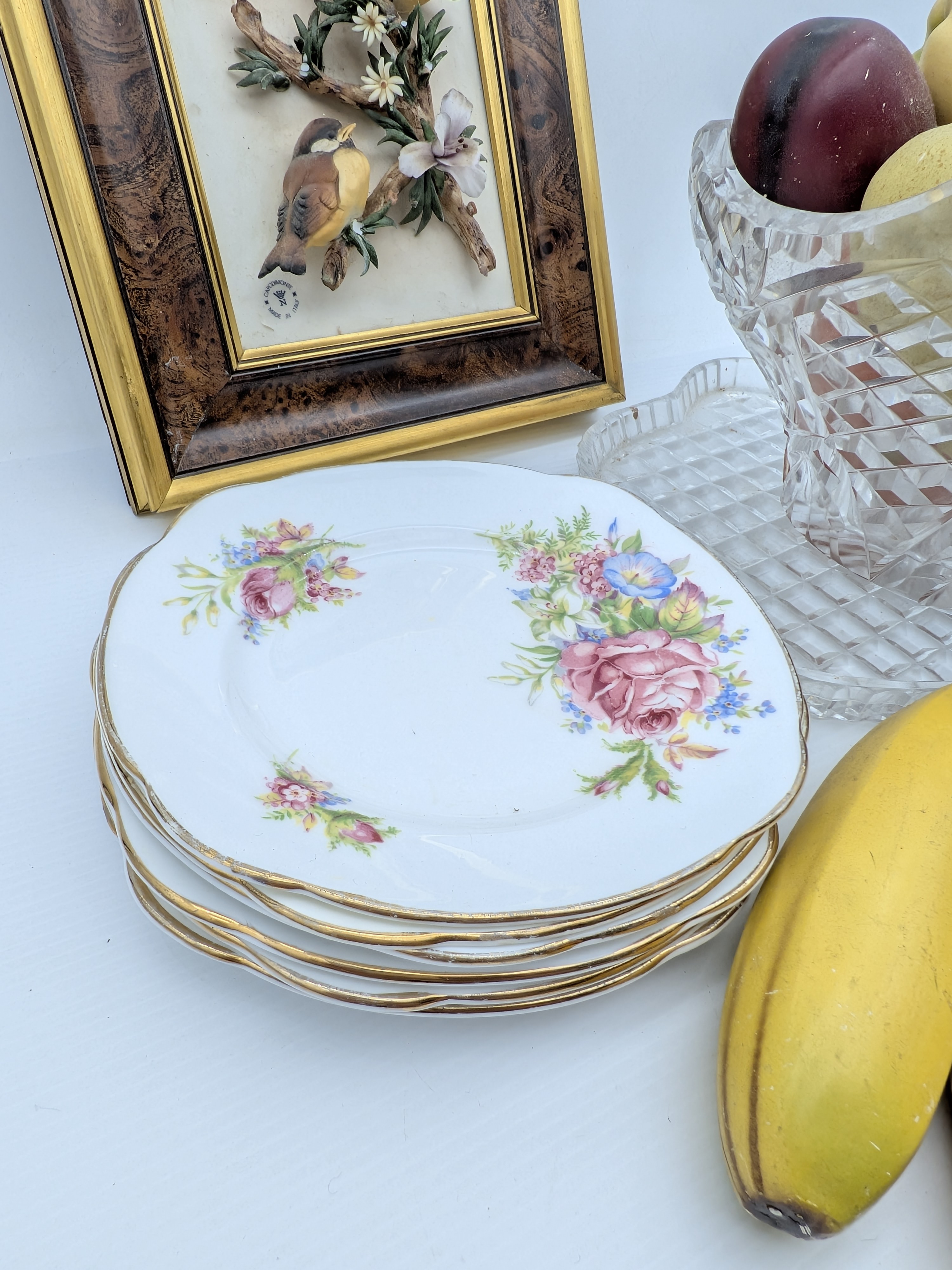 A diverse collection of vintage decorative household sundries, featuring a framed bird relief art piece, a cut-crystal fruit bowl with faux fruit, a stack of ceramic floral side plates, and additional faux fruit. - View 6