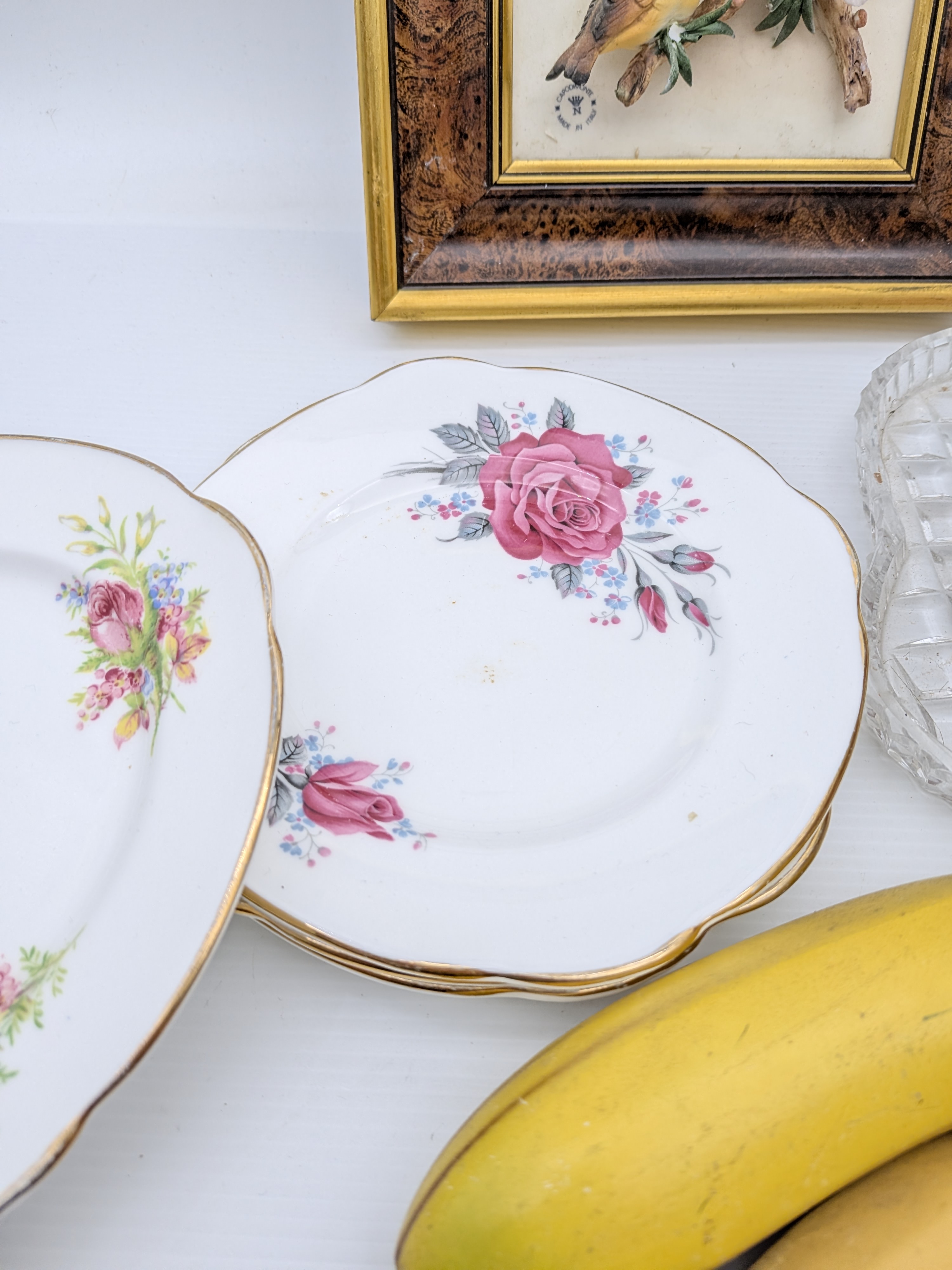 A diverse collection of vintage decorative household sundries, featuring a framed bird relief art piece, a cut-crystal fruit bowl with faux fruit, a stack of ceramic floral side plates, and additional faux fruit. - View 8
