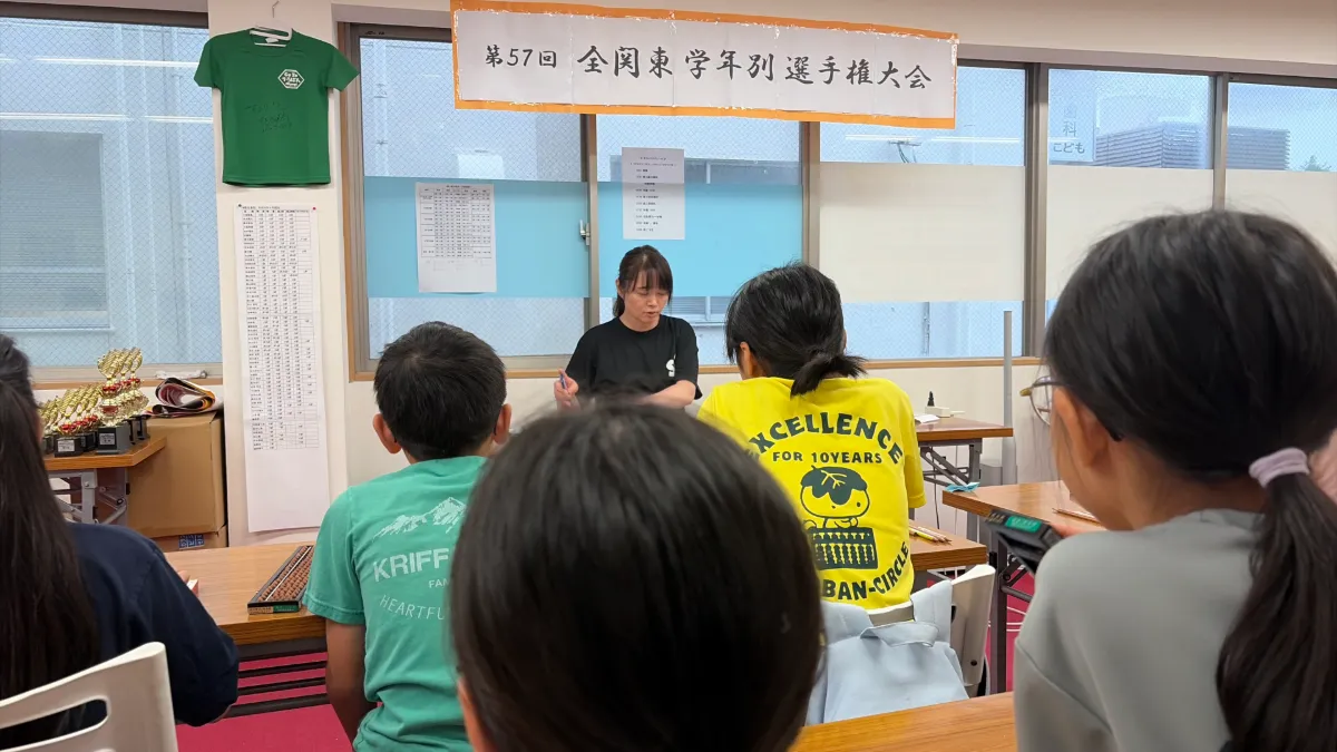 机に向かってそろばんをはじく子どもたち。奥の壁に「第57回 全関東学年別選手権大会」の横断幕が掲げられている