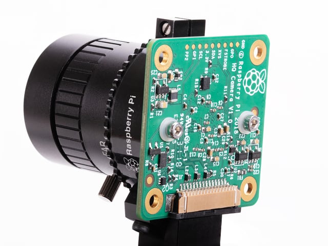 Raspberry Pi camera module with a large lens attached, showing a green circuit board with various components and a ribbon cable connector.