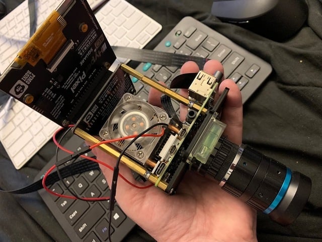 A hand holds a DIY camera setup with a Raspberry Pi 4, a fan, and a lens attached. Wires connect components, with a keyboard in the background. For Low-Light Long-Exposure Photography.
