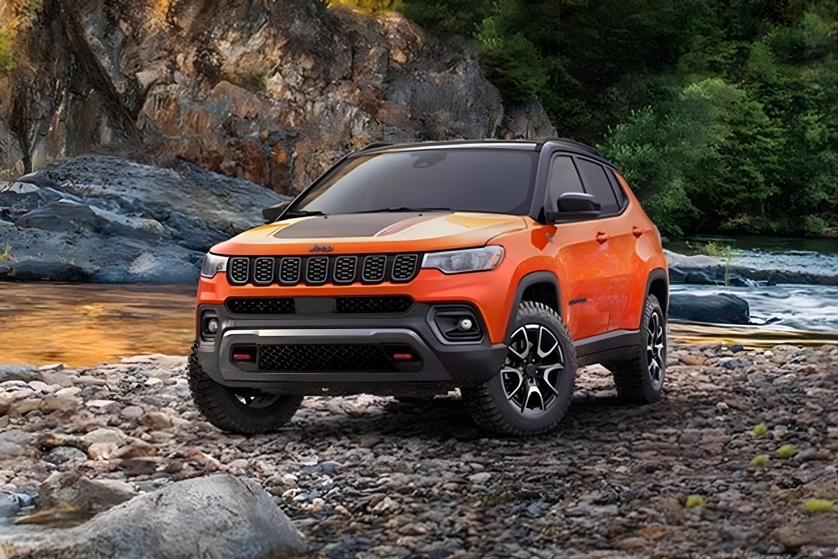 Jeep Compass 2026 orange vu de trois quarts avant stationné sur un terrain rocheux près d’une rivière, mettant en valeur le design robuste et l’aptitude à l’aventure.