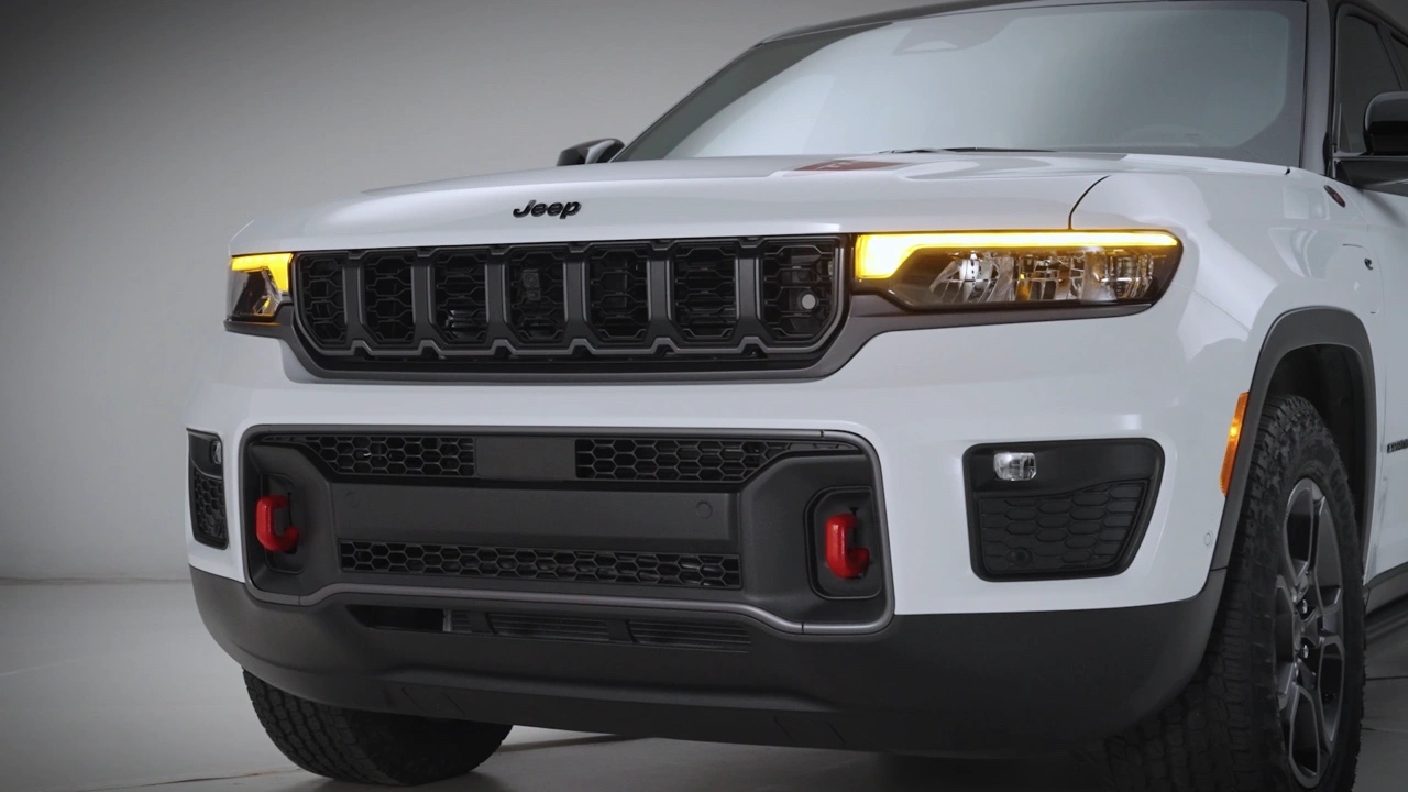 Vue avant du Jeep Grand Cherokee 2026 blanc en studio, mettant en évidence la calandre noire, les feux à DEL et les crochets de remorquage rouges intégrés au pare-chocs.