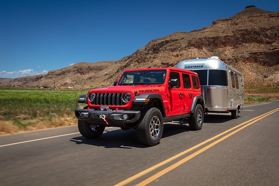Jeep Wrangler Rubicon 2026 rouge remorquant une roulotte Airstream sur route désertique, illustrant la capacité de remorquage, la robustesse du châssis et les aptitudes hors route du VUS.