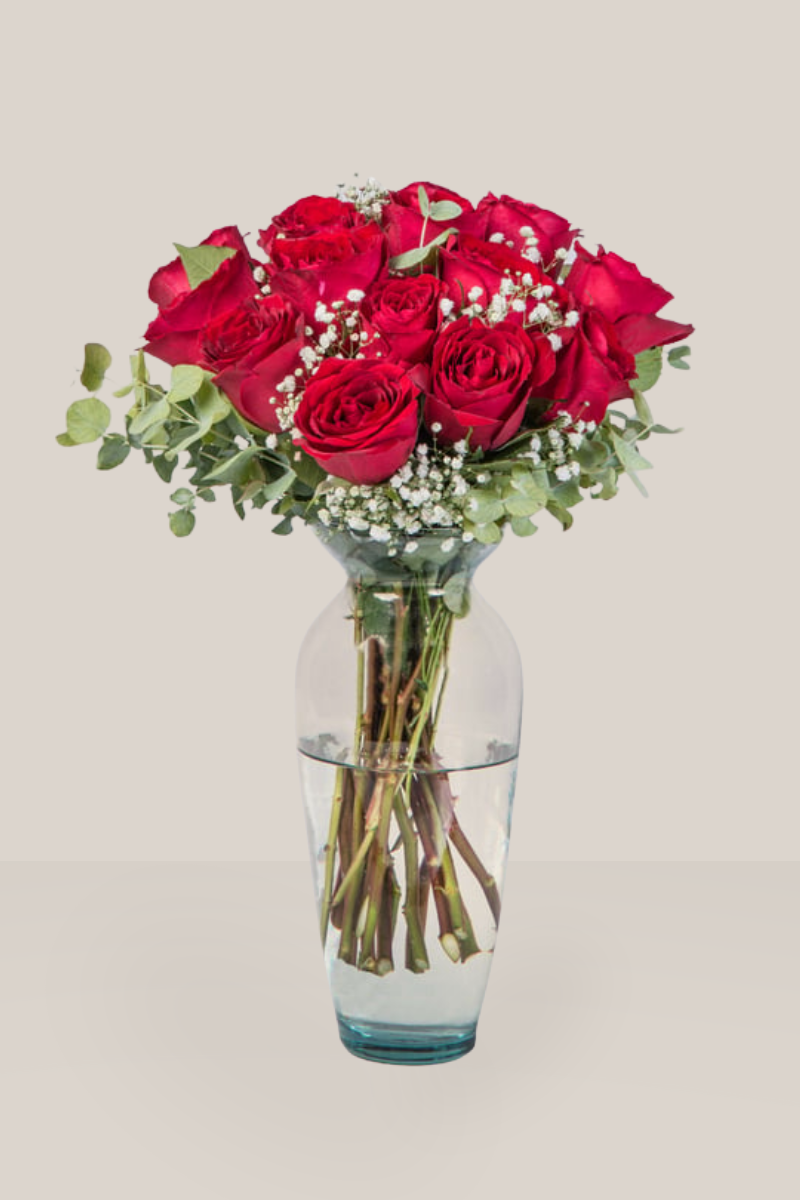 A stunning arrangement of 18 fresh red roses in a clear glass vase, featuring eucalyptus foliage and delicate white baby's breath flowers. The red roses are arranged in a lush, round bouquet. Plain beige background.