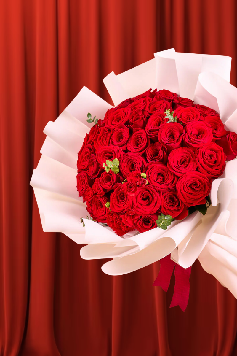 Ramo grande y exuberante de rosas rojas frescas y claveles, con follaje de eucalipto, envuelto en papel blanco y atado con un listón rojo, sobre un fondo de cortina roja.