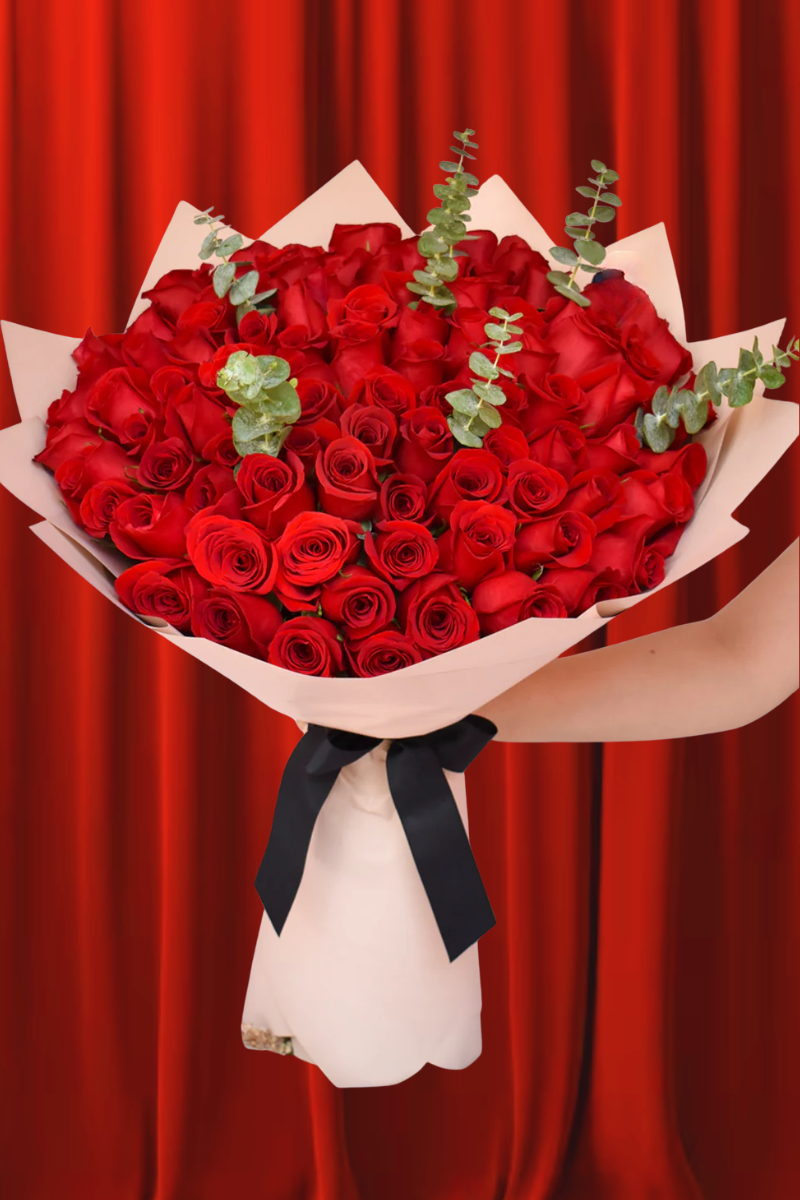 Ramo buchón grande de 100 rosas rojas con tallos de eucalipto, envuelto en papel decorativo beige y atado con un lazo negro, sobre un fondo de cortinas rojas.