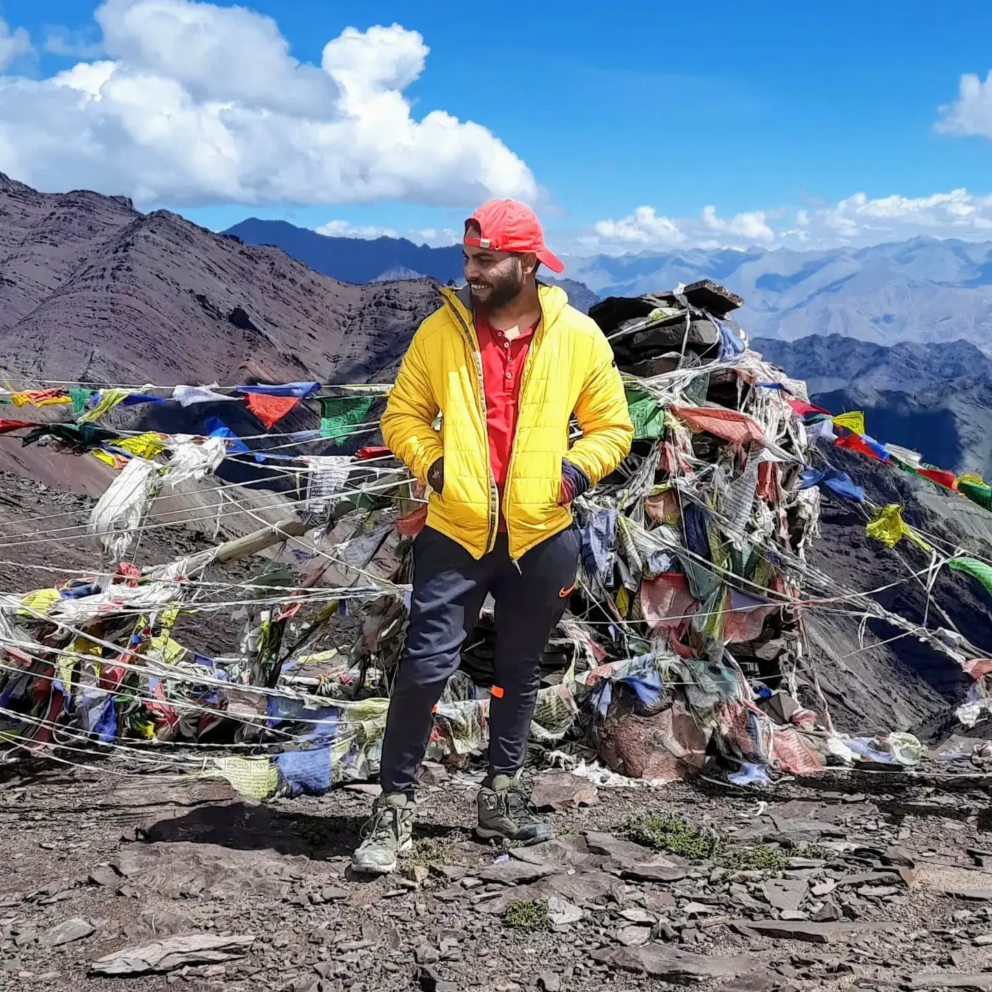 A climber standing at a high-altitude pass surrounded by prayer flags, with rugged Himalayan peaks stretching into the distance under a clear blue sky.
