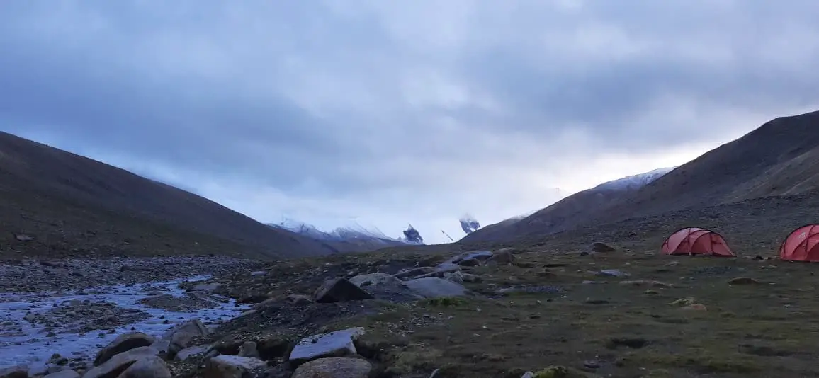 A serene campsite scene at high altitude, with tents pitched beside a rocky valley as clouds roll in over distant snow-covered peaks.