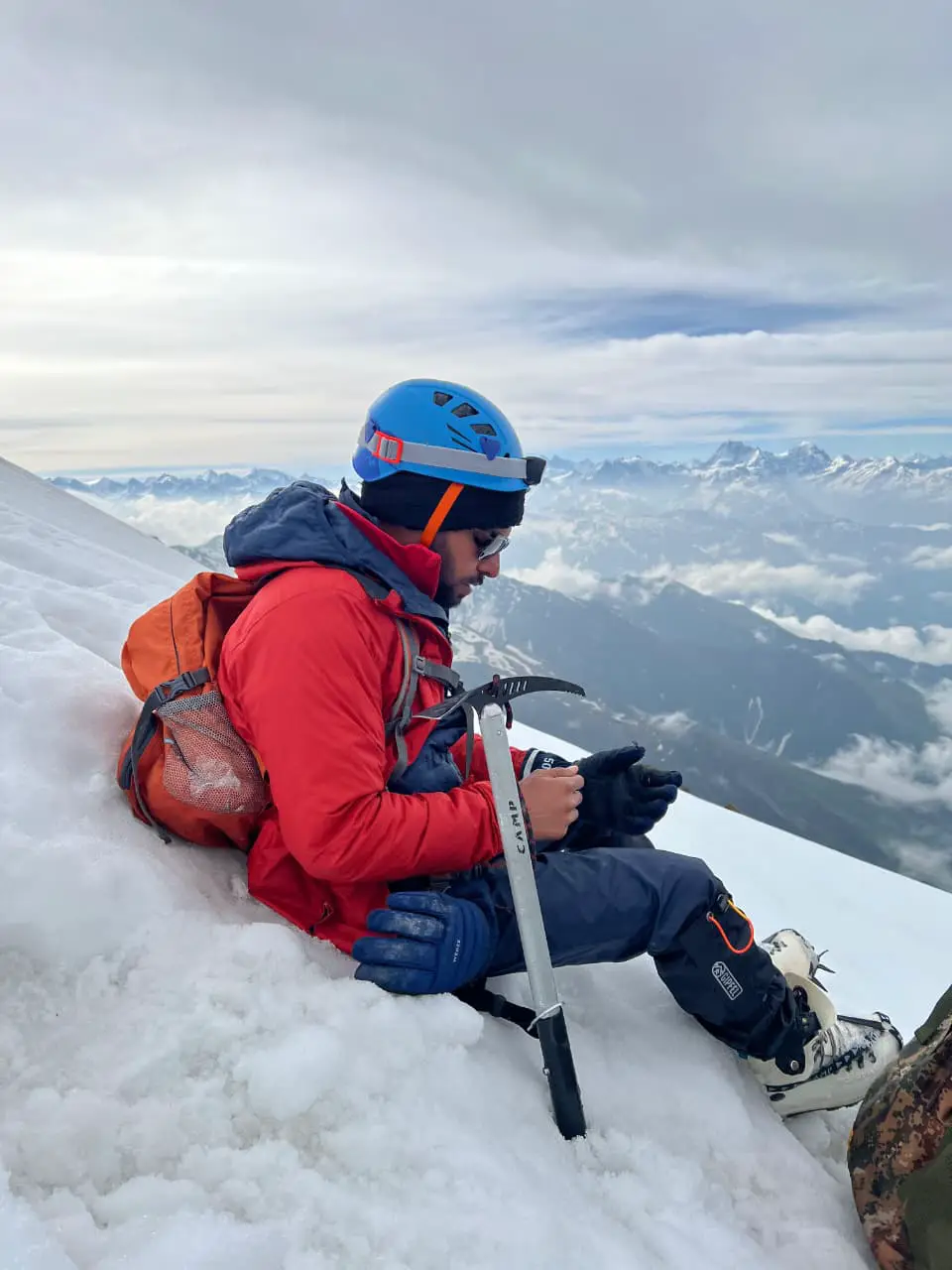 A climber standing at a high-altitude pass surrounded by prayer flags, with rugged Himalayan peaks stretching into the distance under a clear blue sky.