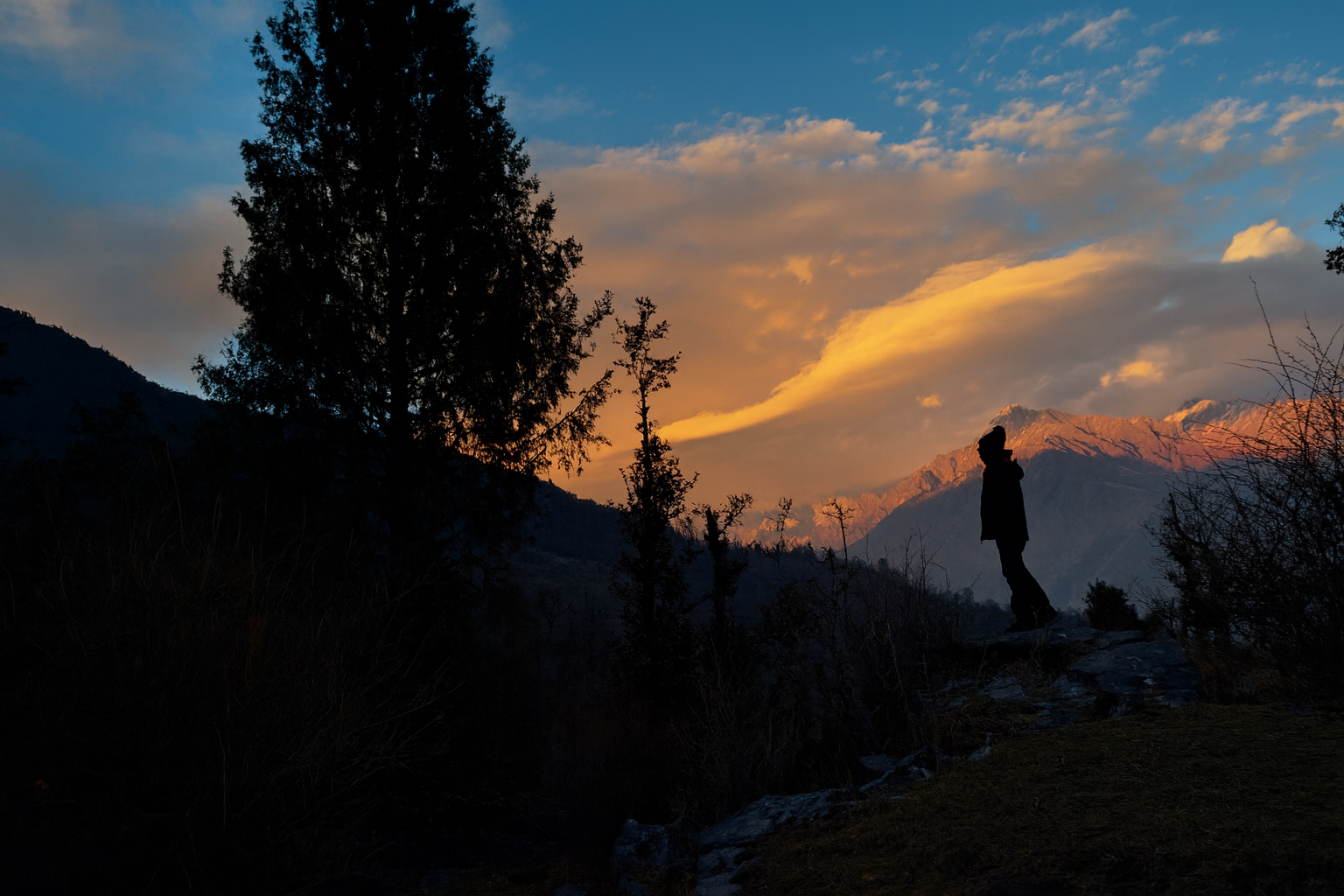 A lone figure faces the mountains at sunrise. The first light turns solitude into peace — a conversation between man and universe.