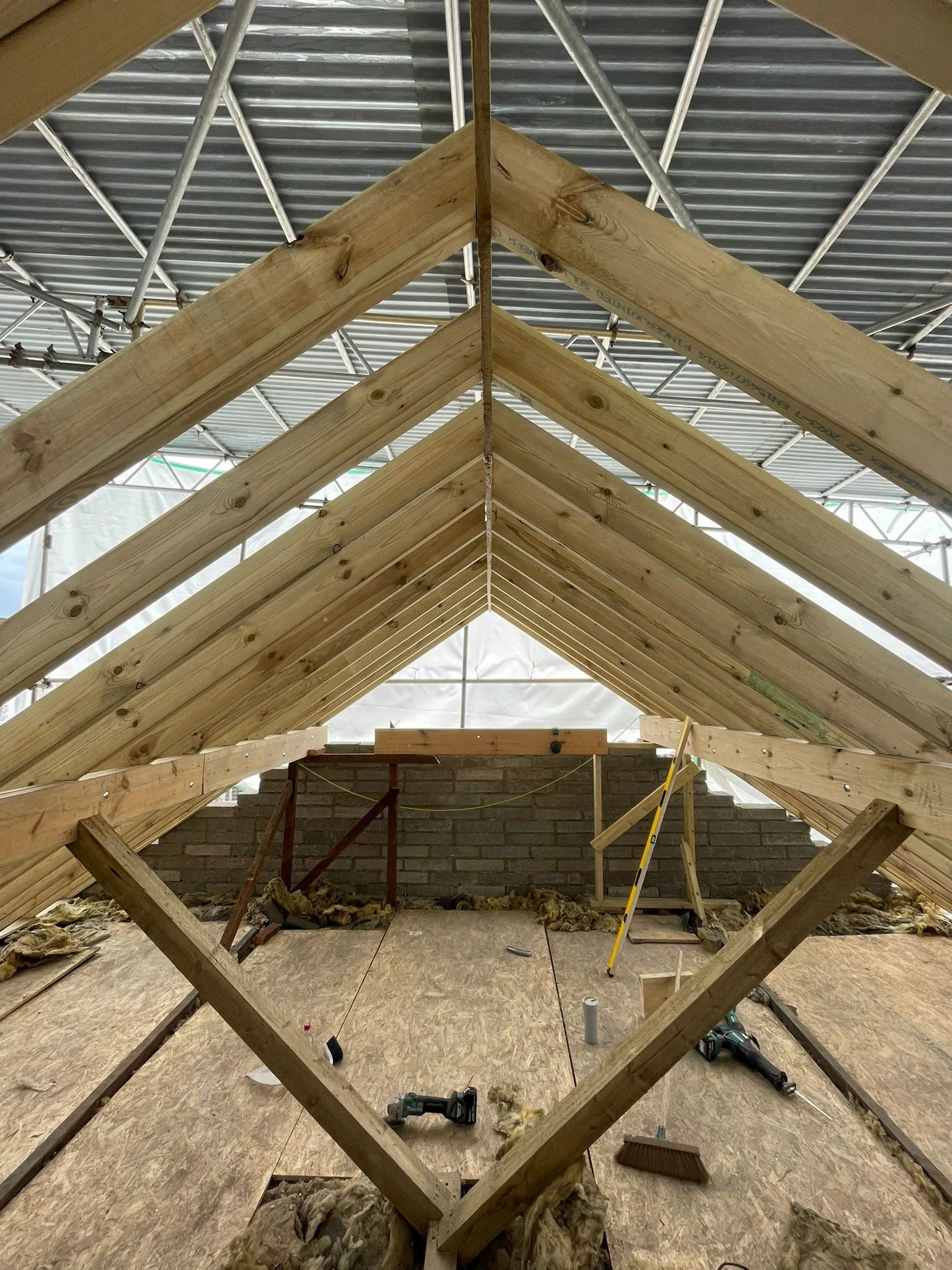 Pairs of rafters meeting at the ridge of a new roof, with diagonal timbers and a boarded deck below, under a scaffold roof