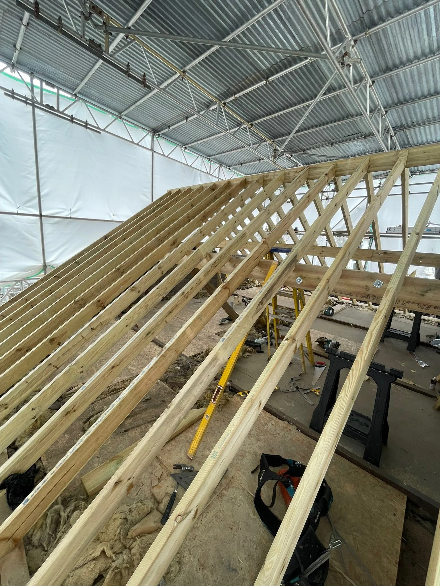 Rafters of a new roof slope with a bolted purlin, sawhorses and carpentry tools on the deck below