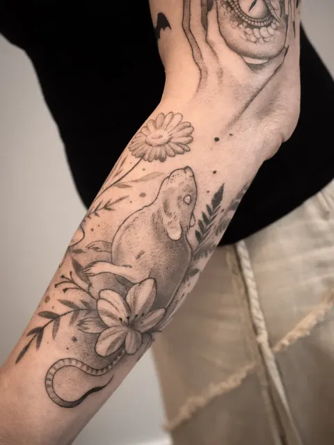 Tatuaż szczura otoczonego kwiatami i liśćmi na przedramieniu, wykonany techniką fineline i dotwork.