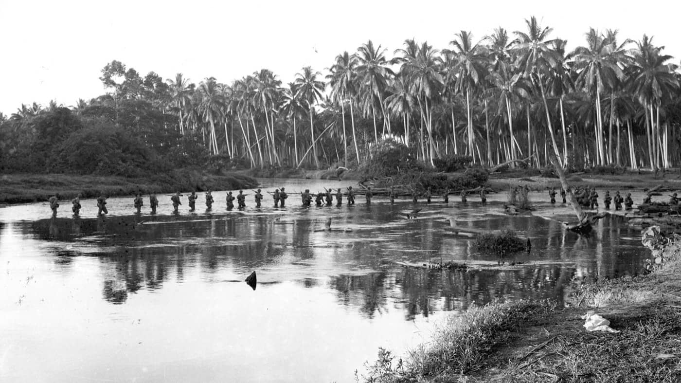 A long line of US Marines wades across a river below coconut palms on Guadalcanal