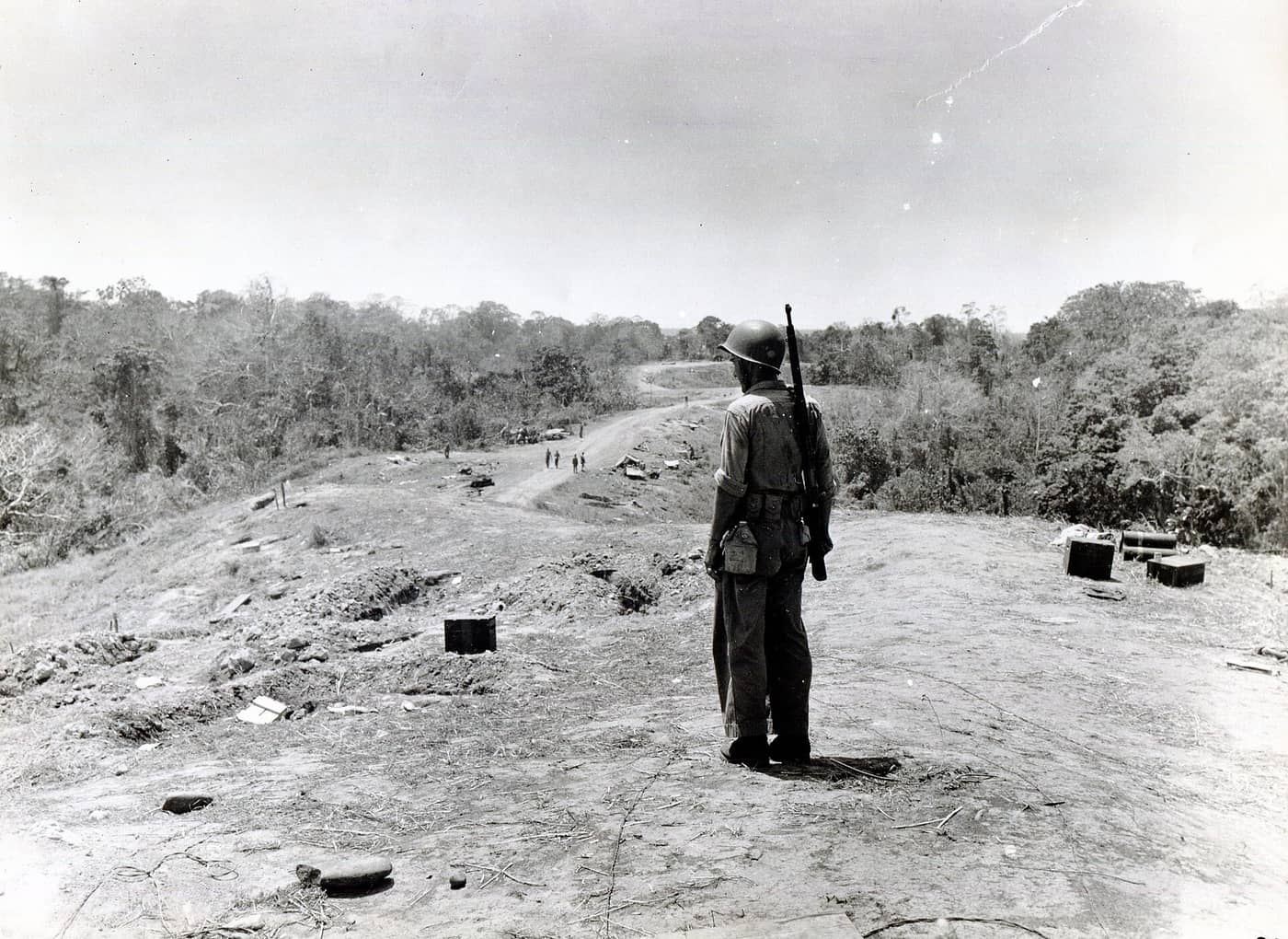A lone Marine with a rifle on his back stands on the bare top of the ridge and looks along it