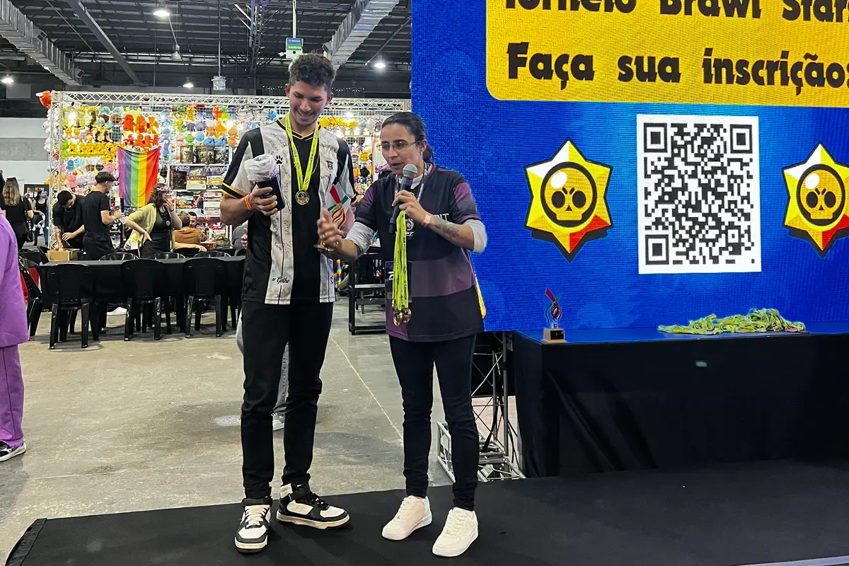 Apresentadores no palco ao lado de um painel com QR Code para inscrição em torneio