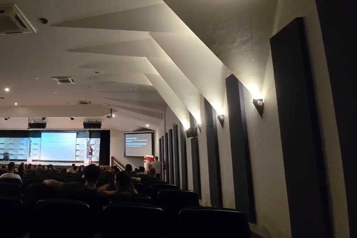Auditório com público assistindo a uma palestra projetada em dois telões
