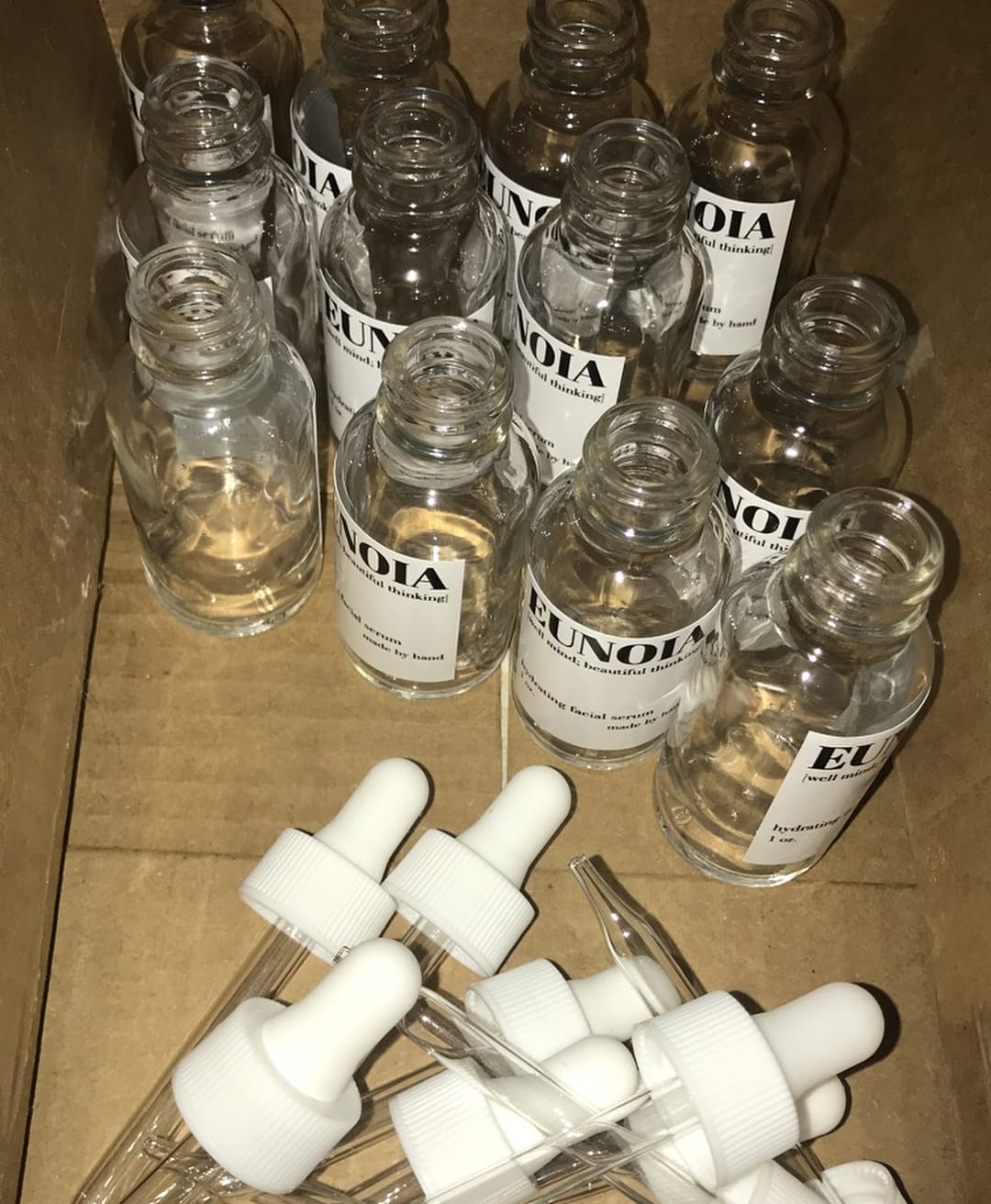 Open bottles of Eunoia's hydrating facial serum beside a pile of droppers in a cardboard box.