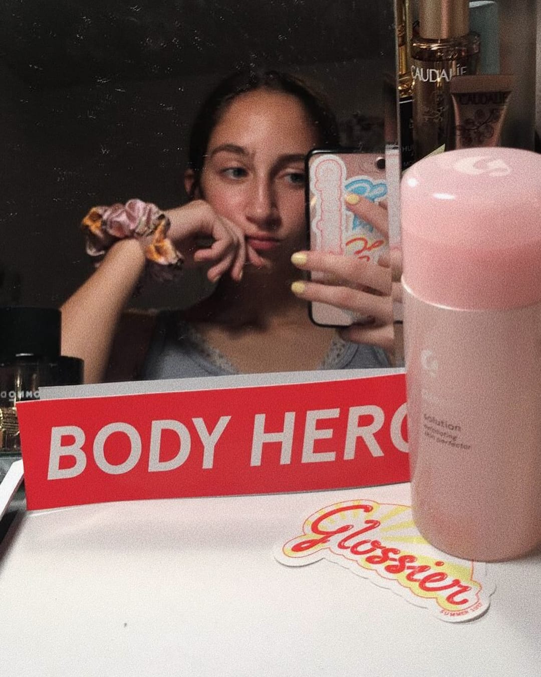 @cafescouture wears a pink scrunchy on her wrist in a mirror selfie with Glossier products and sticker.
