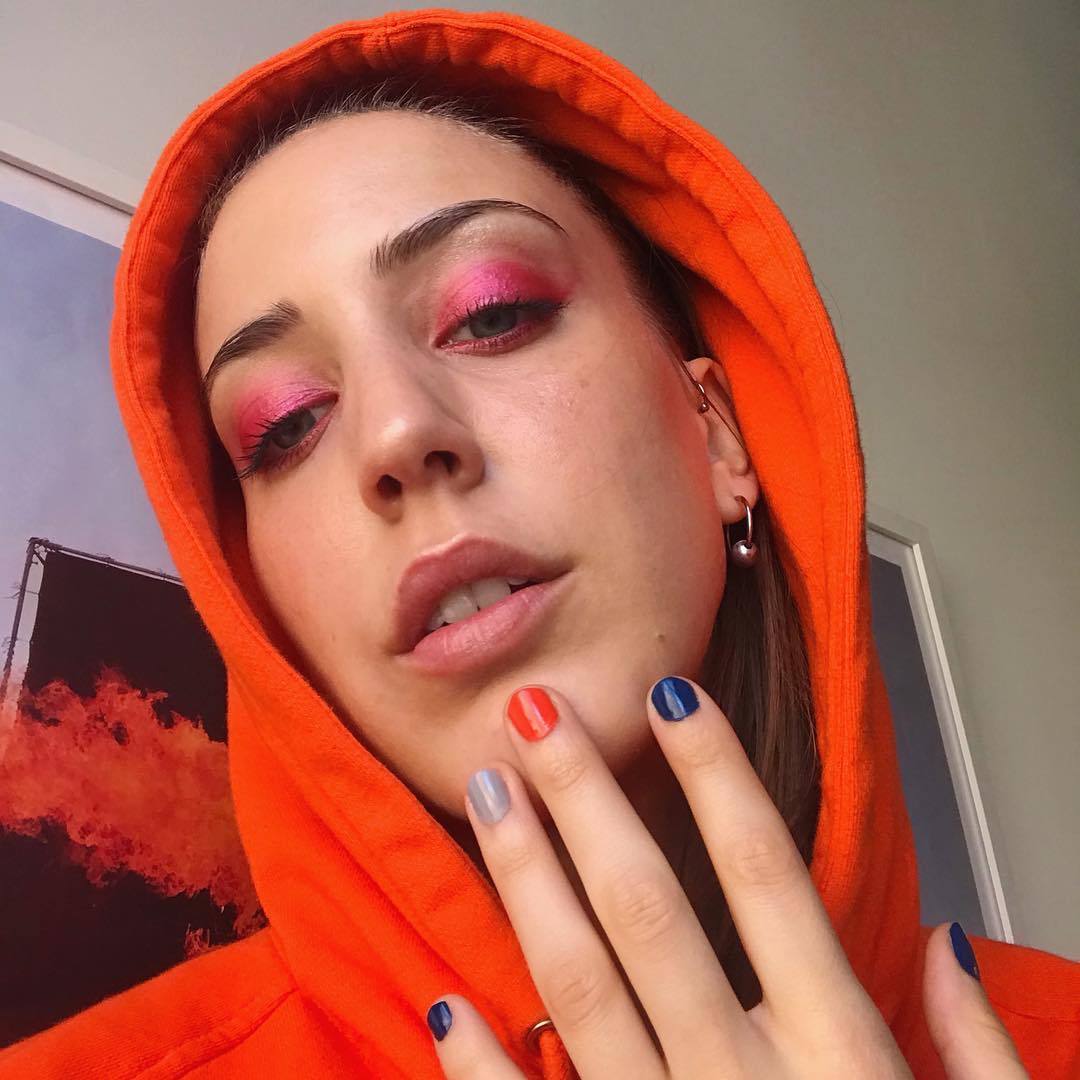 Savannah holds her multicolored nails up to her chin in a selfie where she sports pink eyeshadow and a read hood around her head.