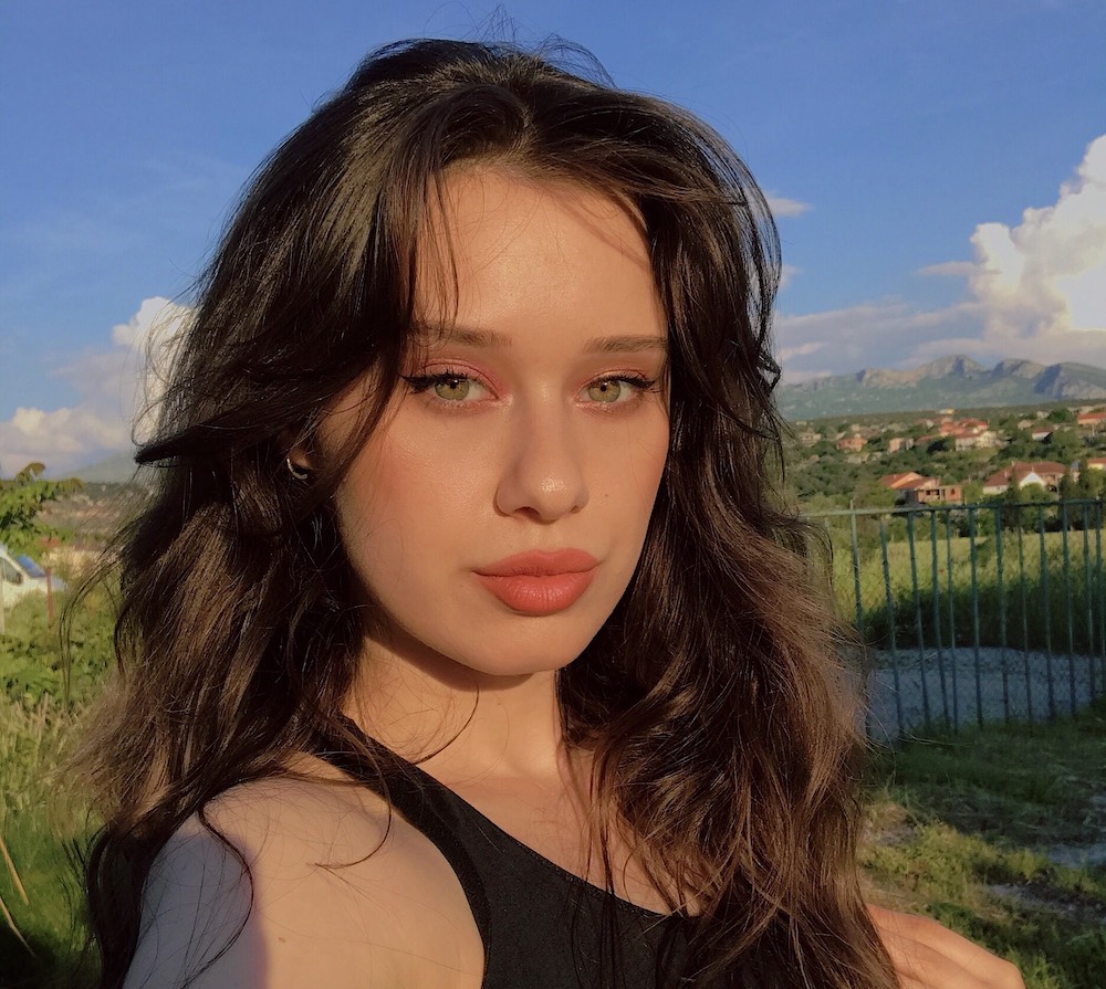 Ajla wears a coral makeup look with voluminous hair in a selfie in front of a luscious green landscape.