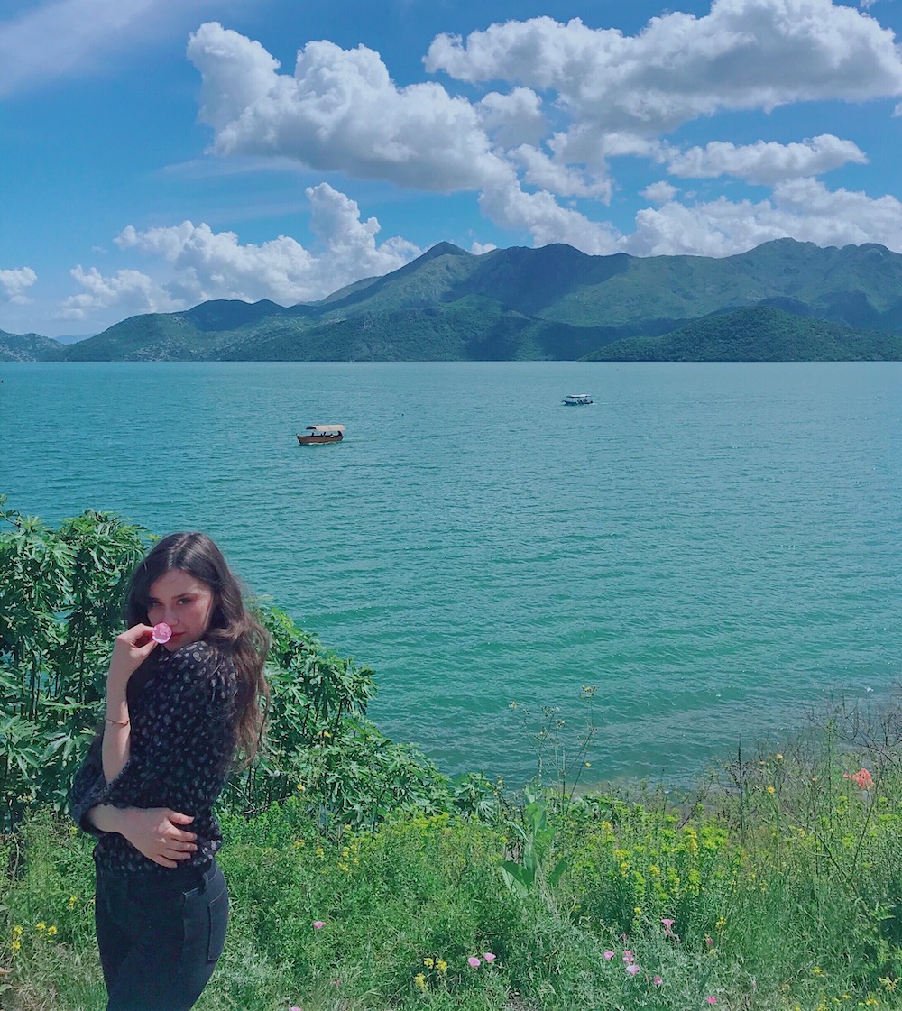 Ajla hols a pink flower to her face as she stands in green grass in front of a teal ocean.