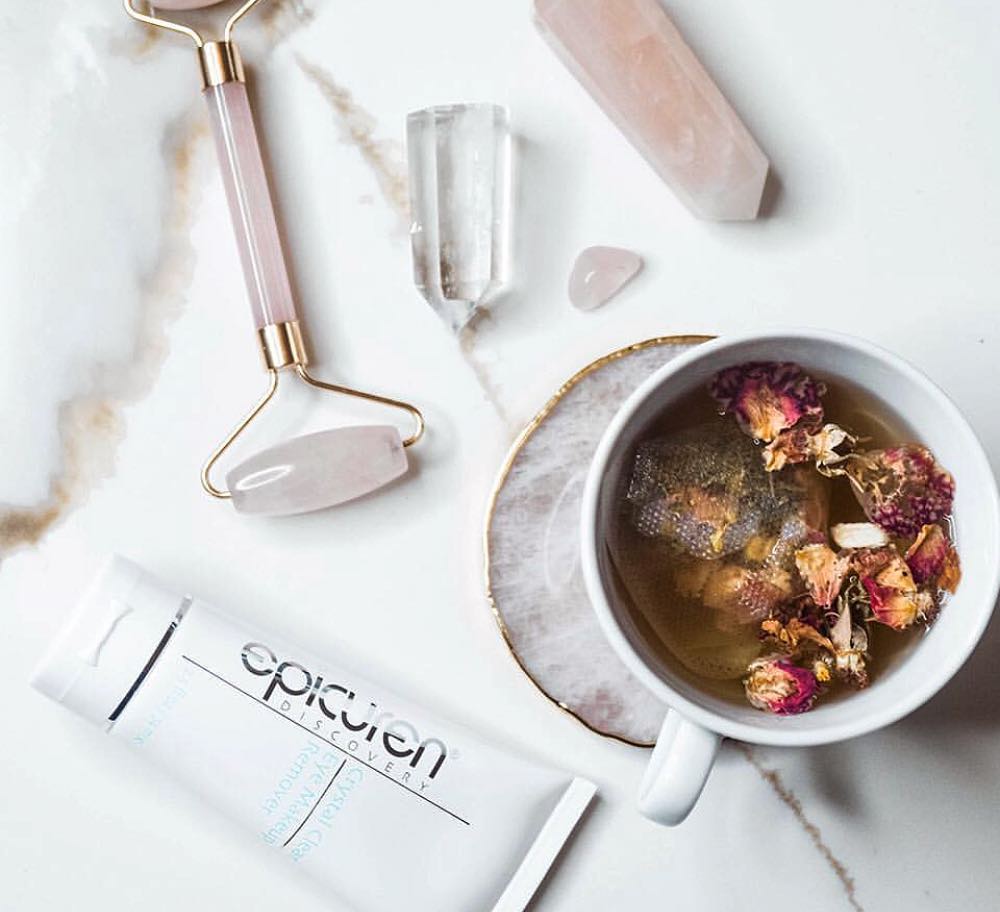 Epicuren's Crystal Clear Eye Makeup Remover layed flat beside a mug of floral tea, a pink jade roller, and three crystals.
