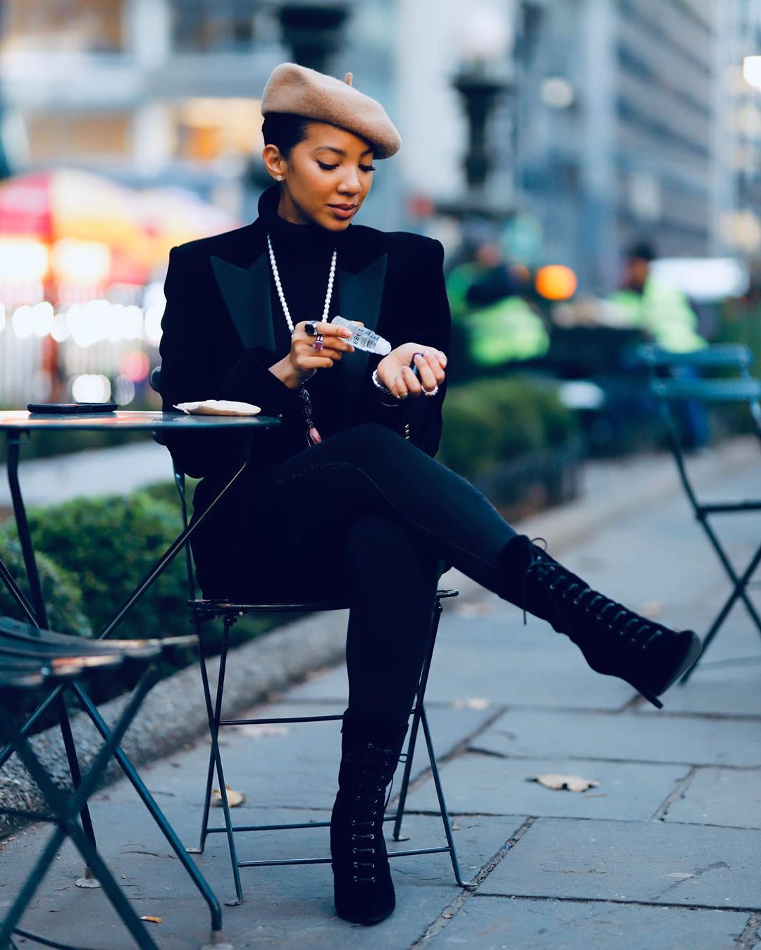 Alyssa Simone applies her perfume while she sits at a cafe-style table and chairs wearing an all-black outfit with a tan beret.