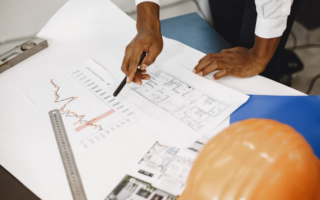 A contractor reviews architectural blueprints and cost charts with a ruler and hard hat nearby, reflecting the disciplined measurement and coordination planning that separates profitable large-job bidding from guesswork.